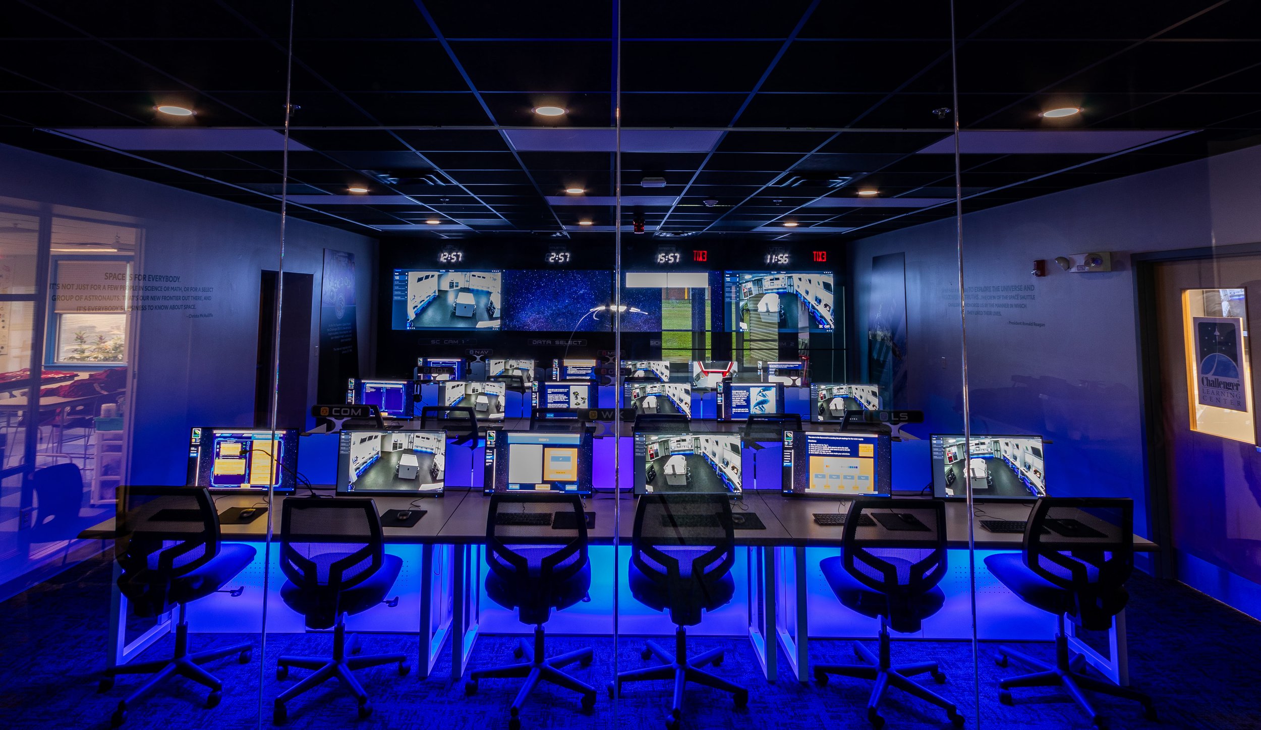 An observation room with multiple computer stations and monitors showing a space-themed simulation or control panel, illuminated with blue lighting.