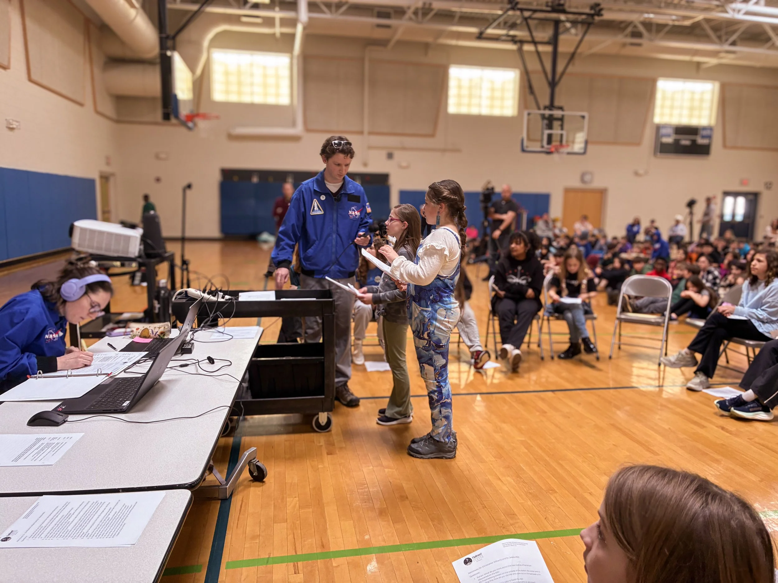 Children and adults participating in a science or space-themed educational event in a gymnasium. Kids are presenting projects or speeches to an audience sitting on chairs.
