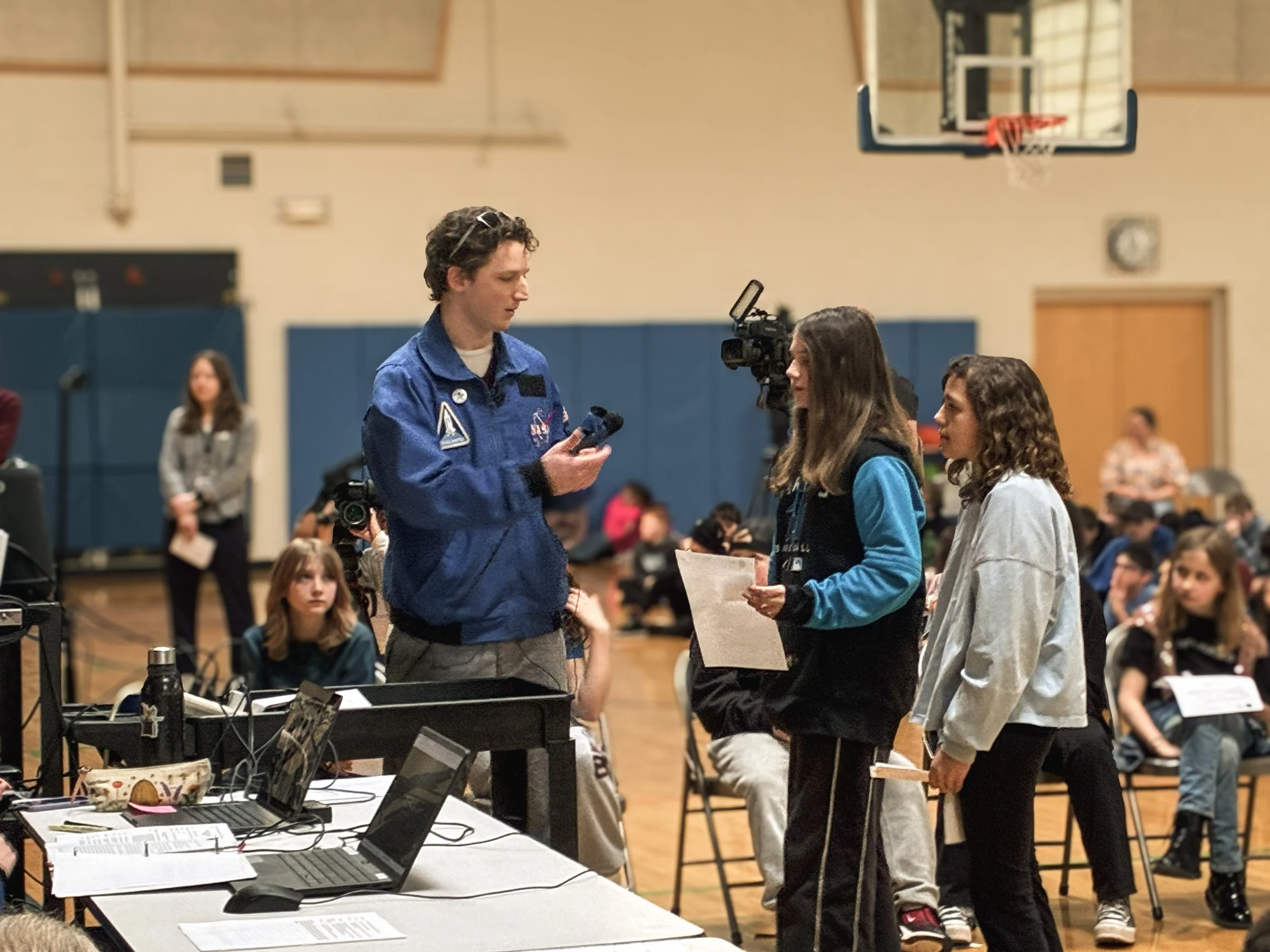 A man in a blue NASA jacket speaking to two young girls holding papers in a gymnasium or auditorium with other children seated in the background. A camera on a tripod is filming the interaction.
