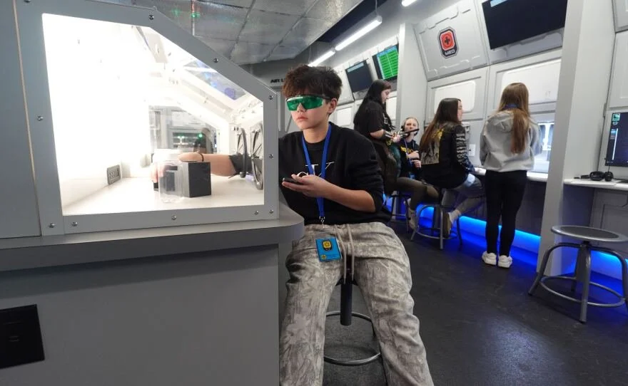 A young person wearing green-tinted glasses and casual clothing sitting at a science exhibit, with other kids working at computer stations in the background inside a high-tech facility.