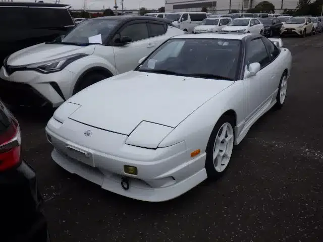 White Nissan 240SX sports car parked at a car dealership or lot, surrounded by other vehicles.