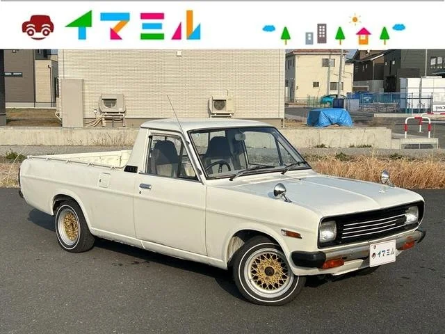 A vintage white pickup truck with gold wheels parked on a street, with residential buildings and blue sky in the background.
