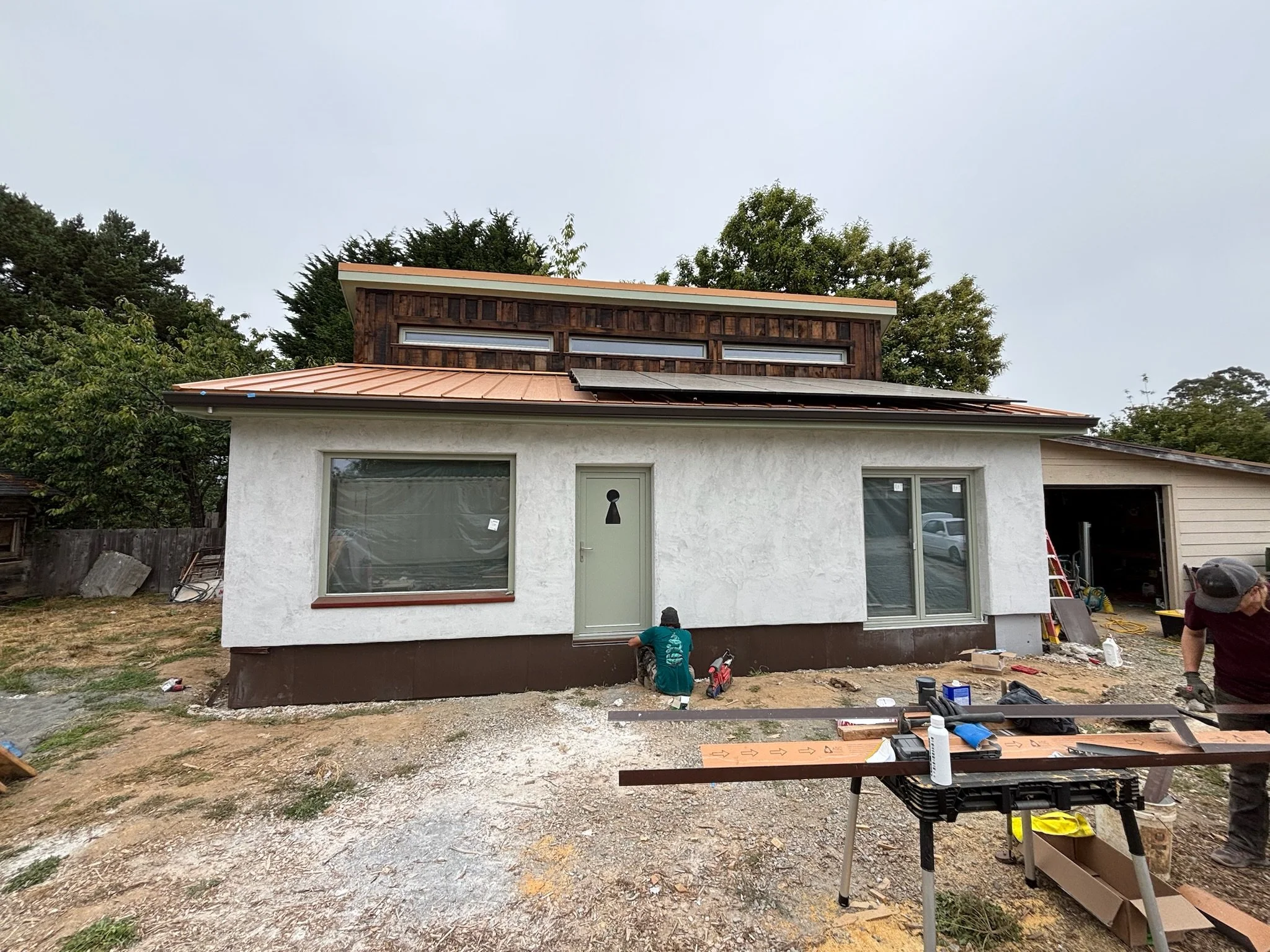 House under construction with workers installing siding and trim, in a yard with tools and materials.