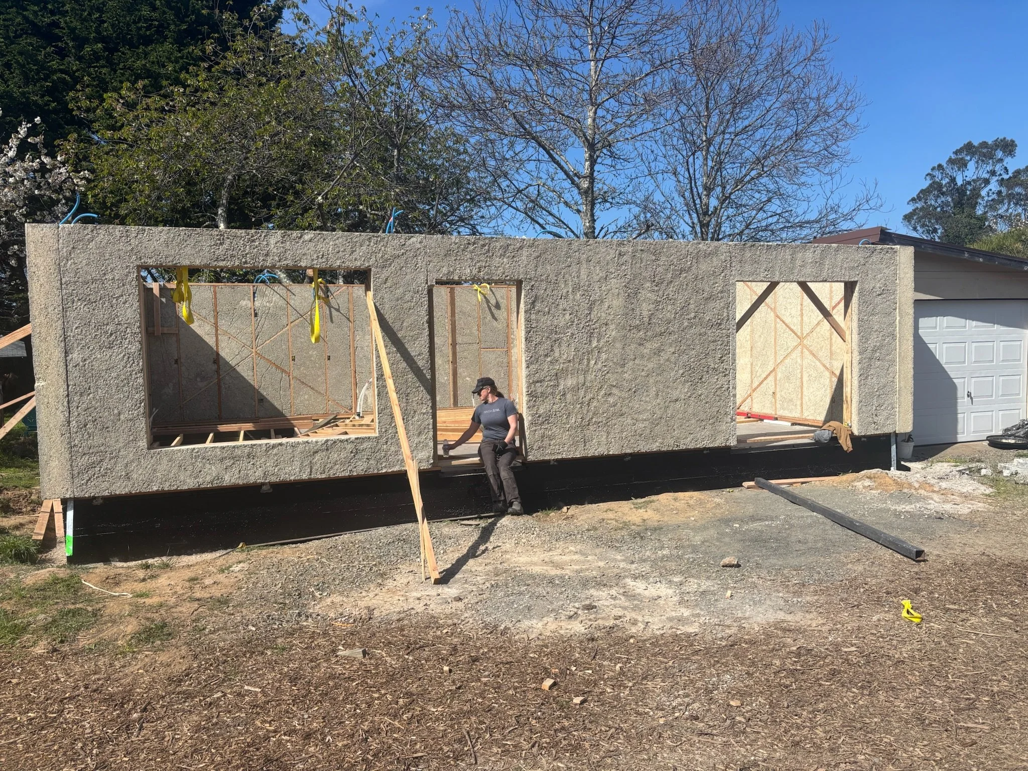 Construction of a new building with exterior walls partially completed, showing wooden framing and insulation, with a person sitting on the edge of the foundation near the garage.