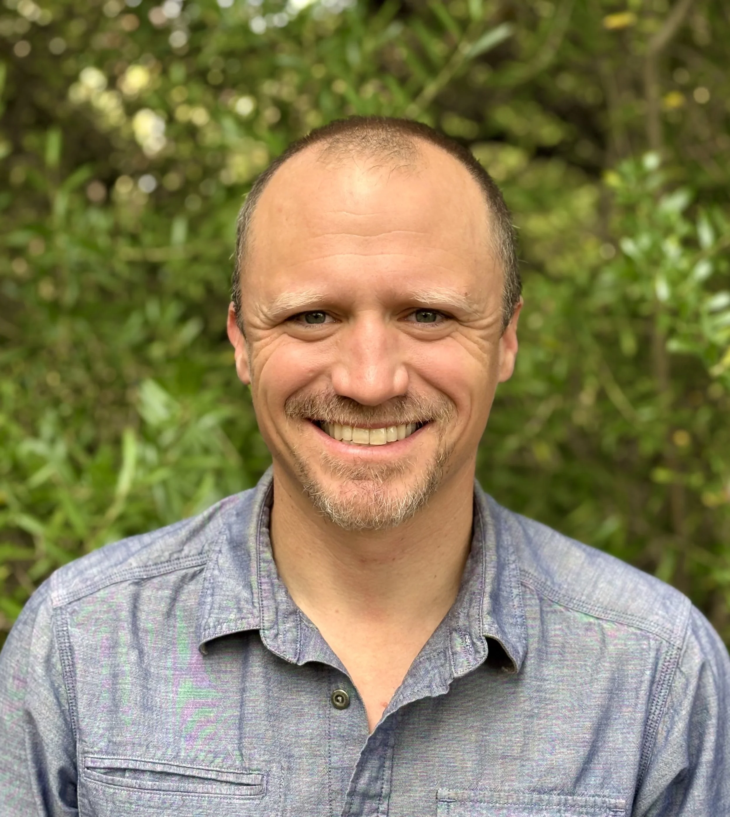 A man smiling outdoors with a body of water and cliffs in the background, wearing a maroon cap.