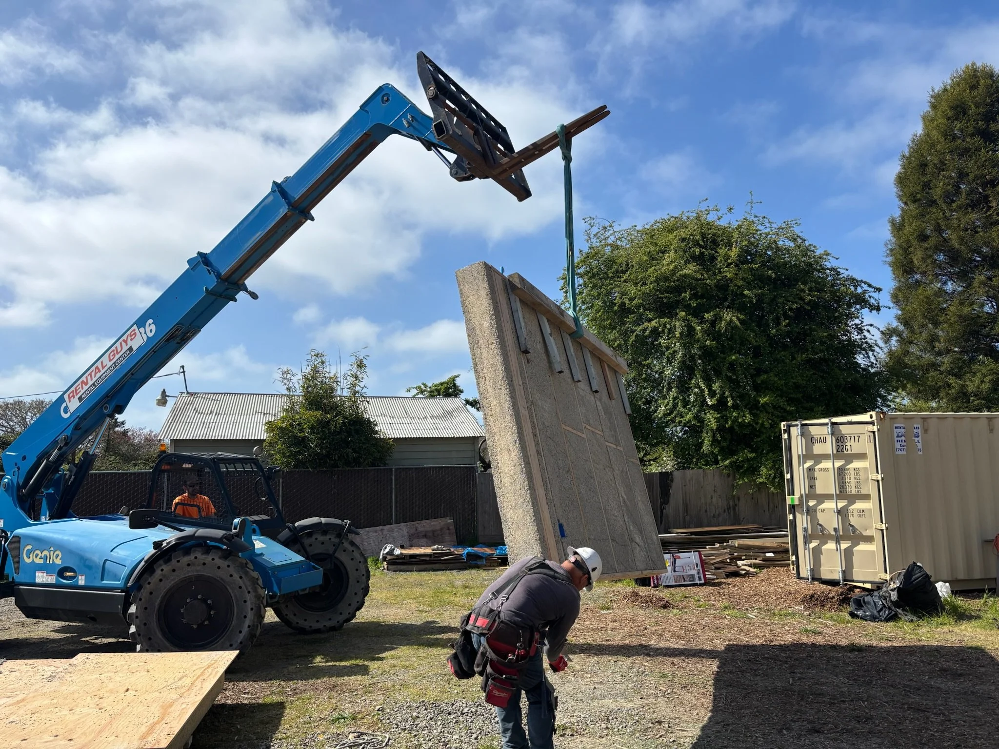 A construction worker is working on a construction site with a blue crane lifting a large concrete panel. The worker wears a white safety helmet and safety harness, and there is a person in an orange safety vest in the background near a black fence. A container and a wooden structure are also visible.