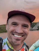 A man smiling outdoors with a body of water and cliffs in the background, wearing a maroon cap.