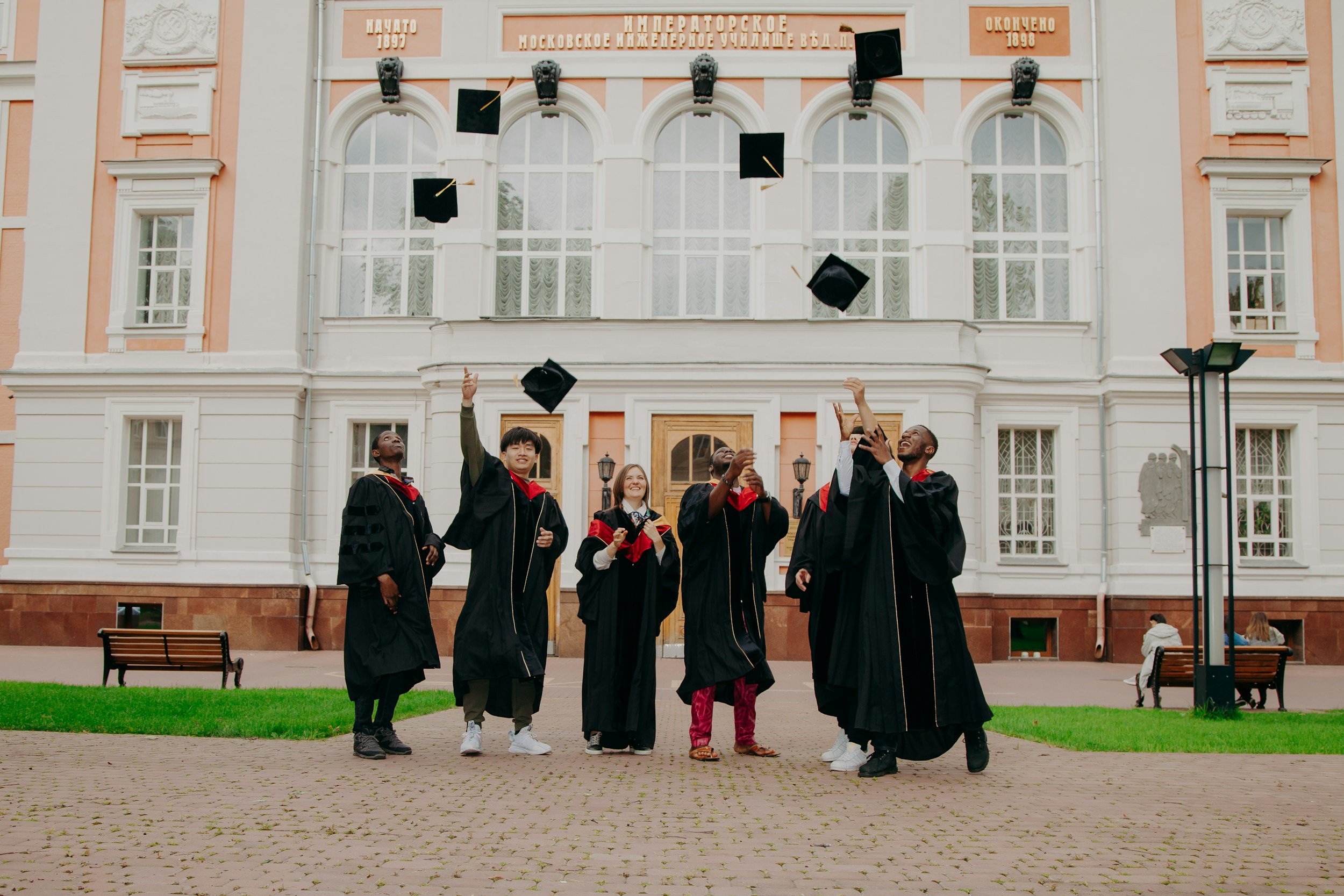 Group of diverse students in graduation gowns celebrating outdoors in front of a historic building, tossing graduation caps into the air.
