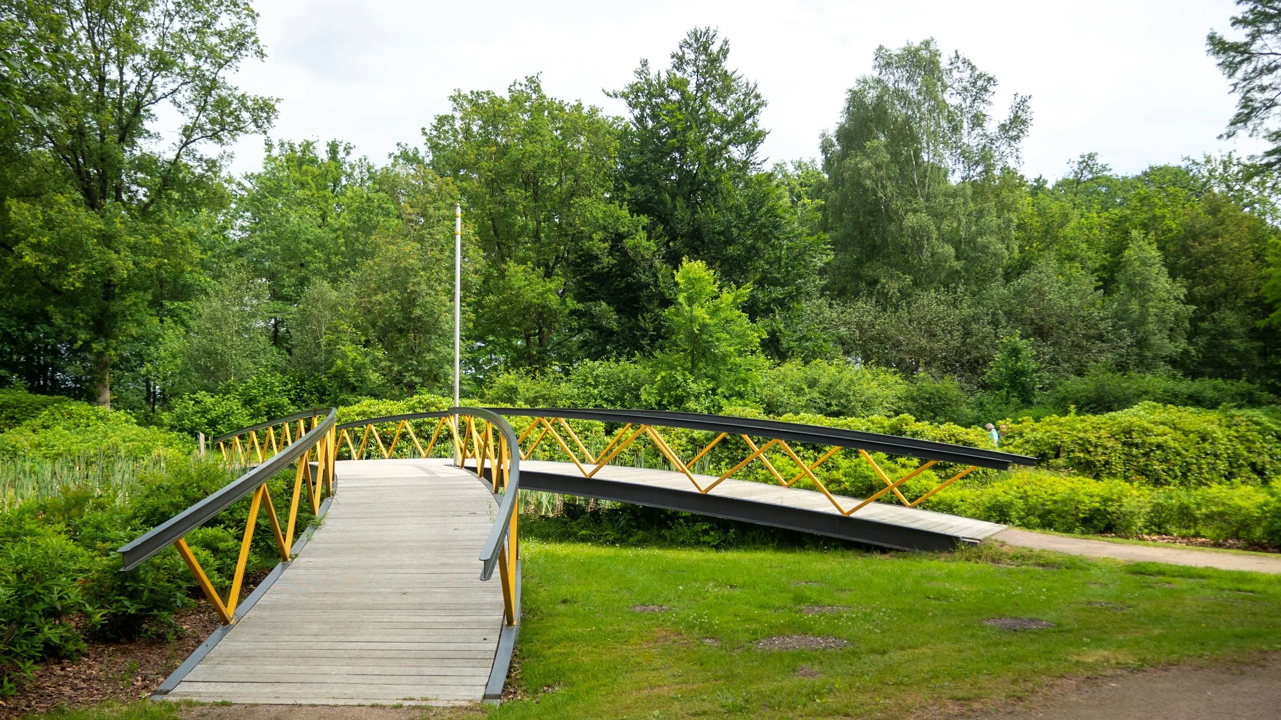 A small curved footbridge with yellow railings over a grassy area, leading into a lush green park with trees.