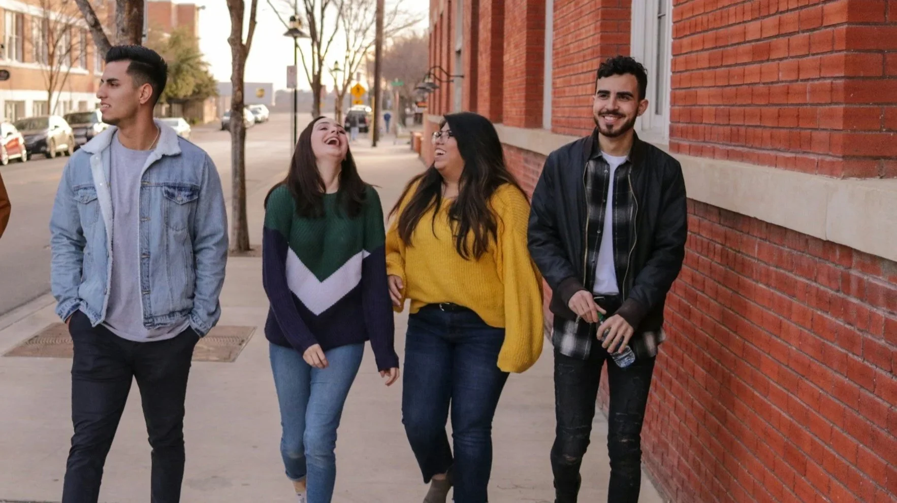 Four friends walking along a sidewalk, smiling and laughing, near a brick building with trees in the background.