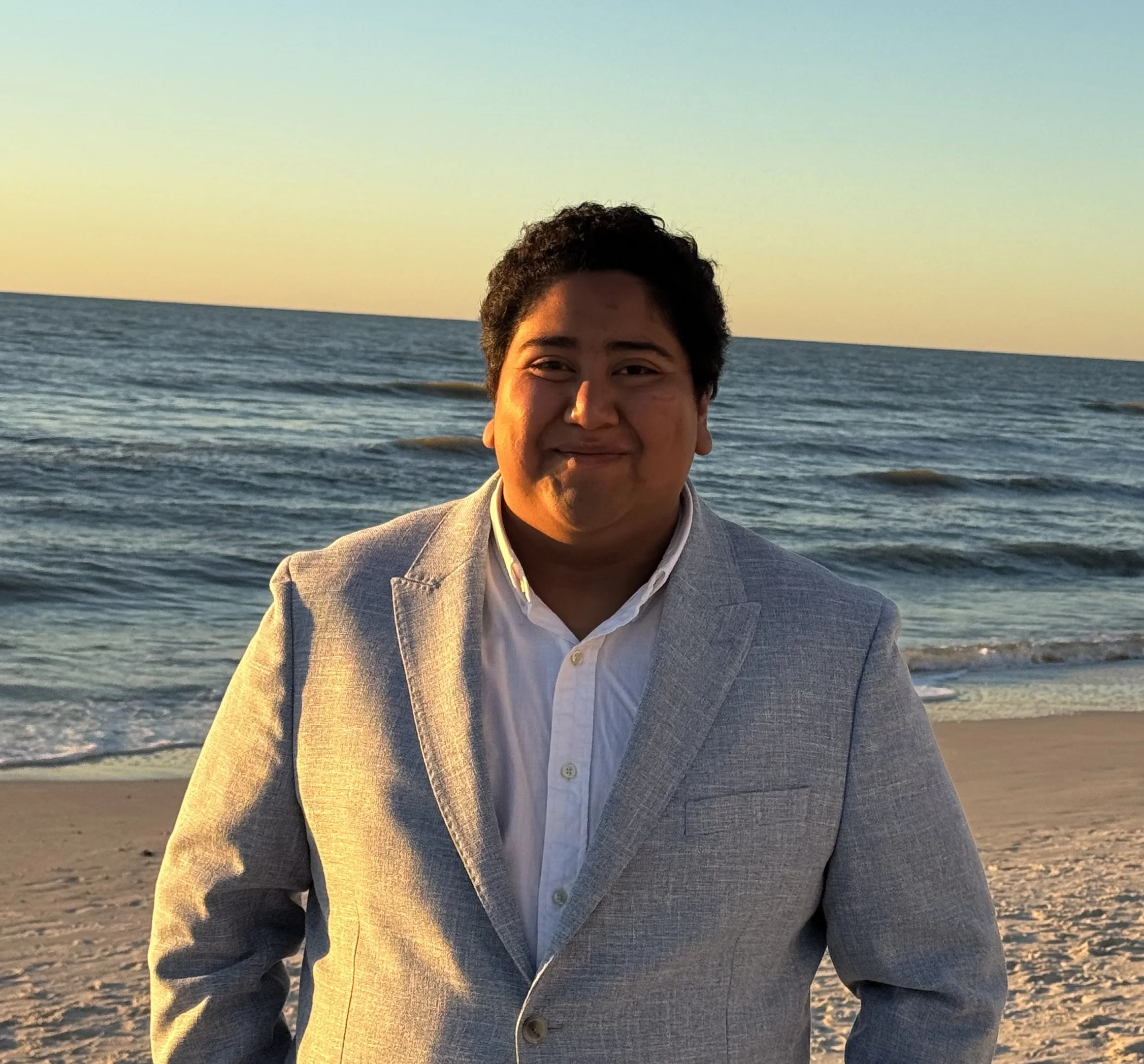 A man in a light gray suit jacket and white shirt standing on a beach at sunset.