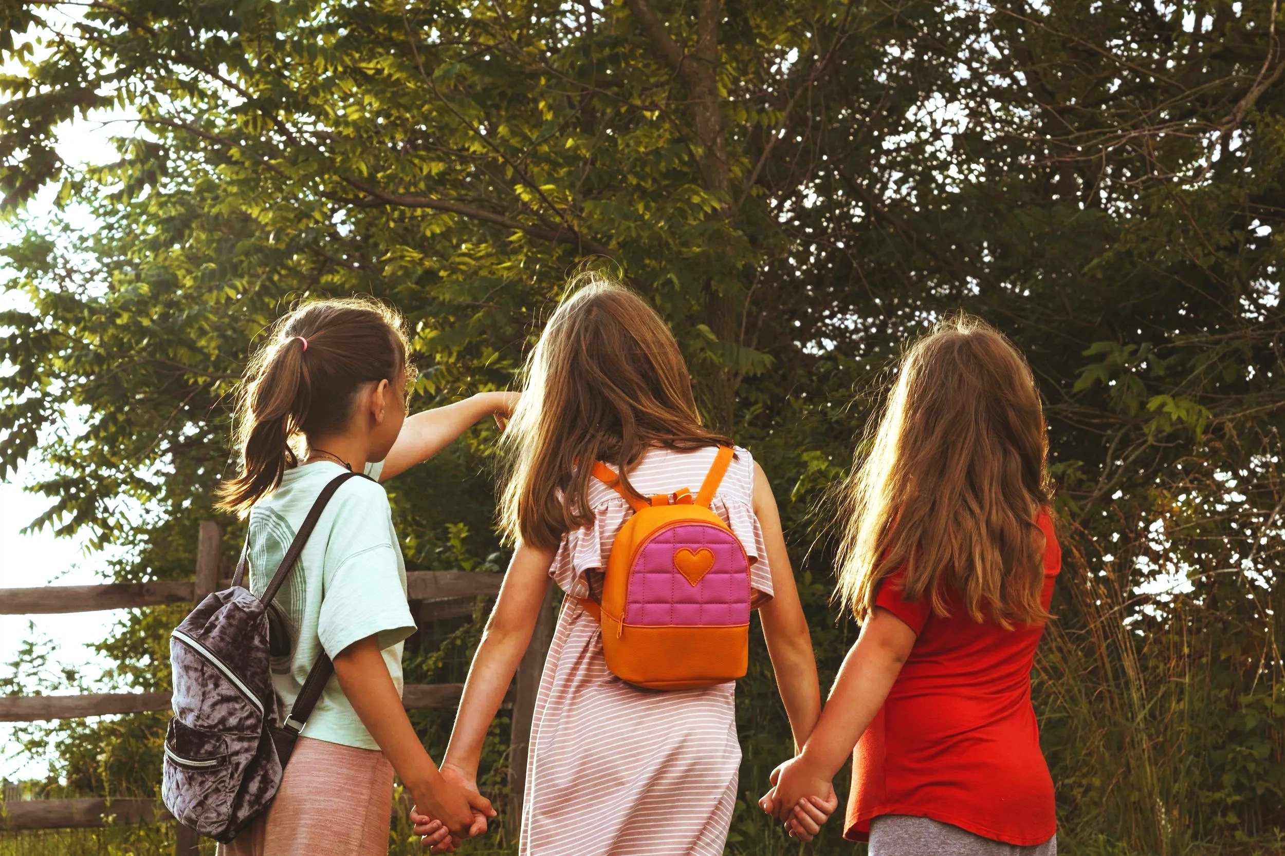 Three young girls holding hands in a circle outdoors, wearing colorful backpacks, near a wooden fence and trees, during daytime.
