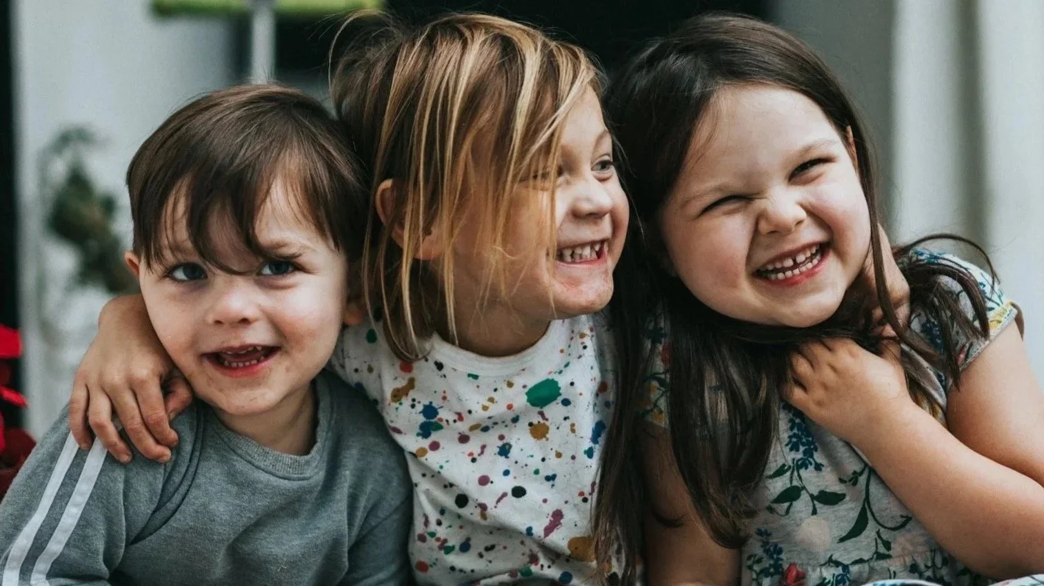 Three young children smiling and laughing together, with their arms around each other.