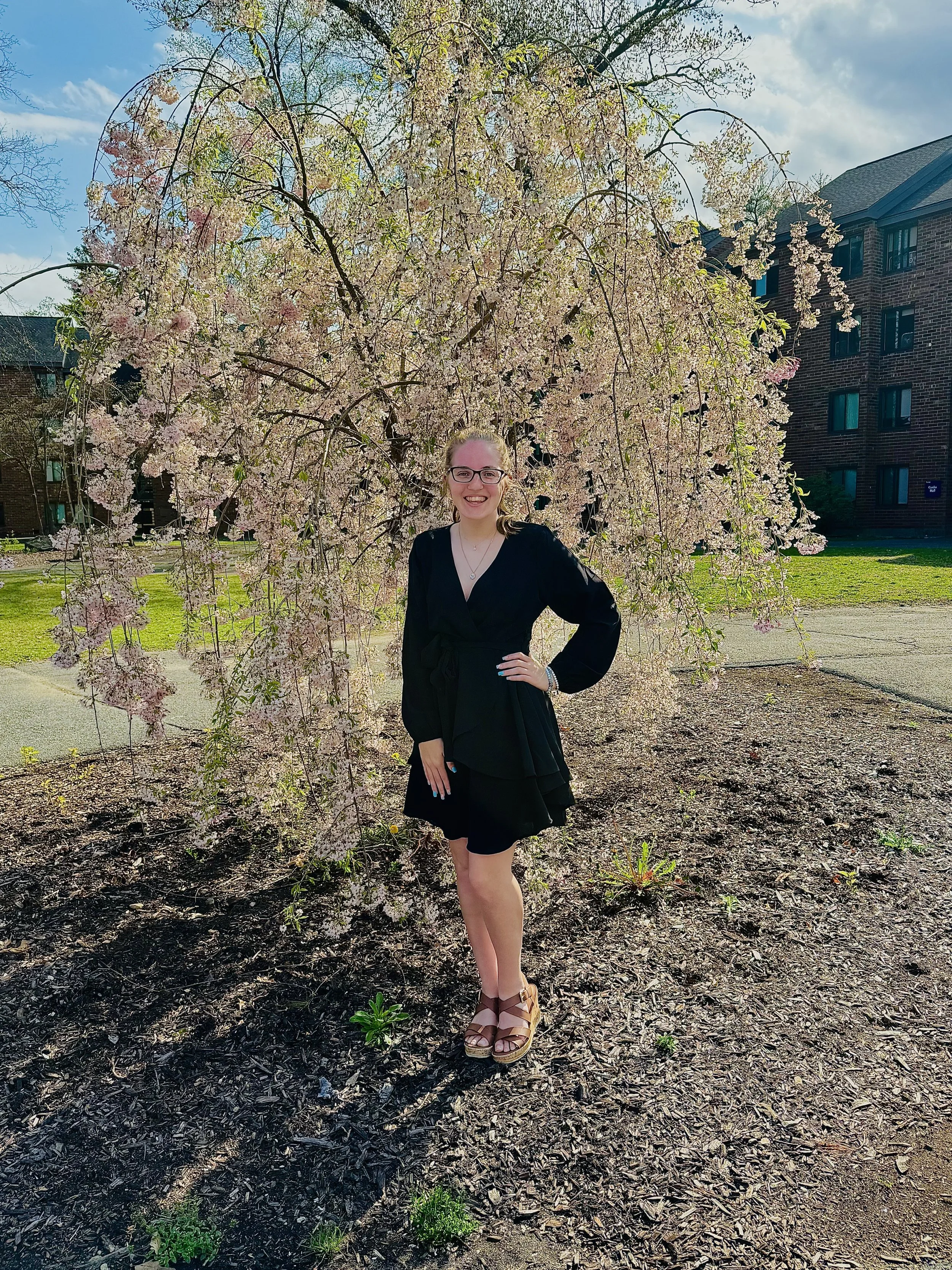 A woman in a black dress and tan sandals standing in front of a blooming cherry blossom tree, with apartment buildings in the background on a sunny day.