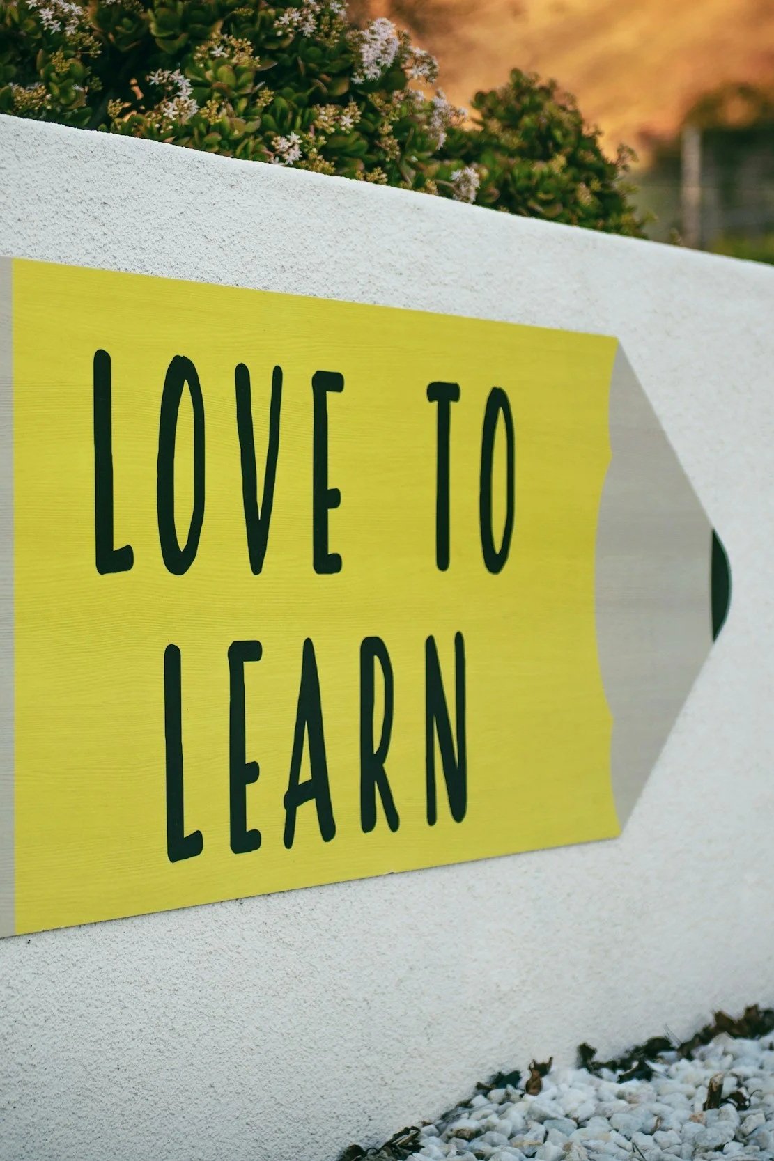 A yellow sign with black text that reads 'Love to Learn' attached to a white wall outdoors.