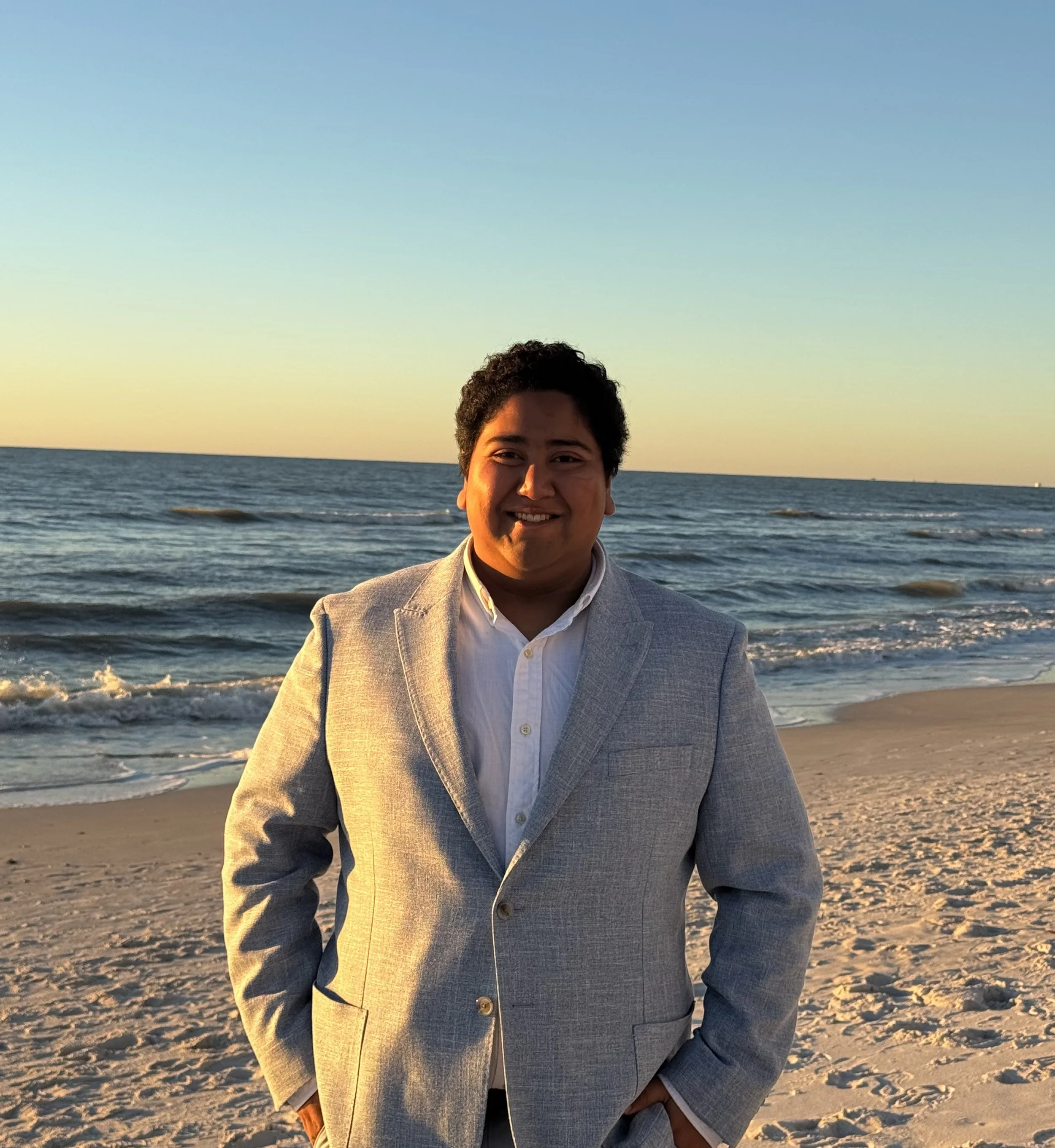 A smiling man in a light gray blazer and white shirt standing on a sandy beach during sunset, with the ocean and a clear sky in the background.