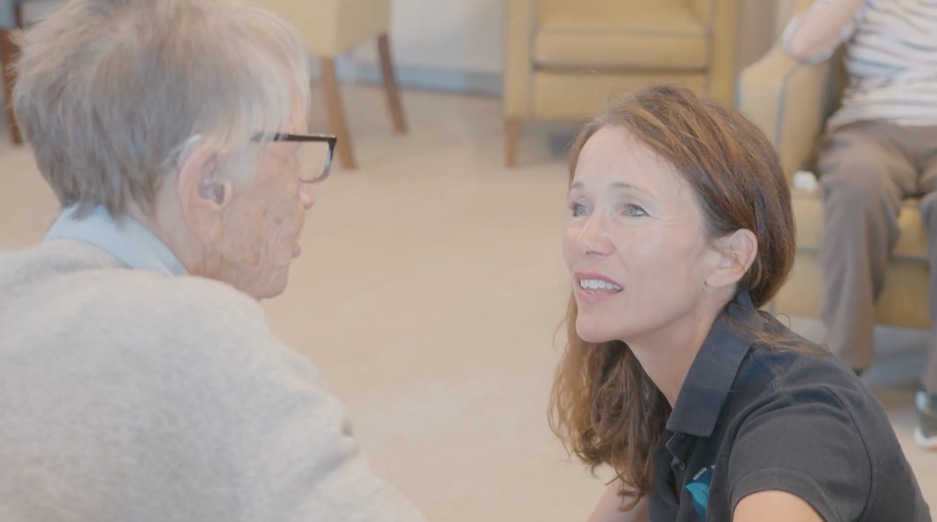 A woman with brown hair and a black shirt looking up emotionally at an older man with gray hair and glasses, sitting face to face indoors.