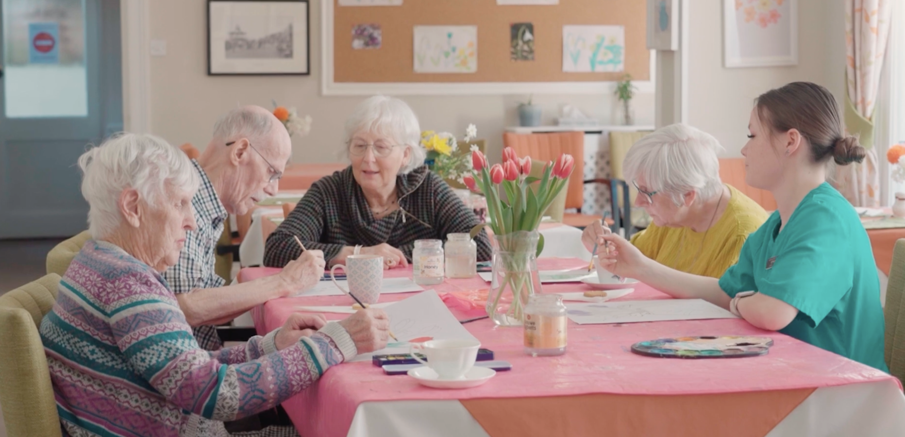 Elderly people and a caregiver engaging in a painting activity at a table decorated with flowers in a community room.