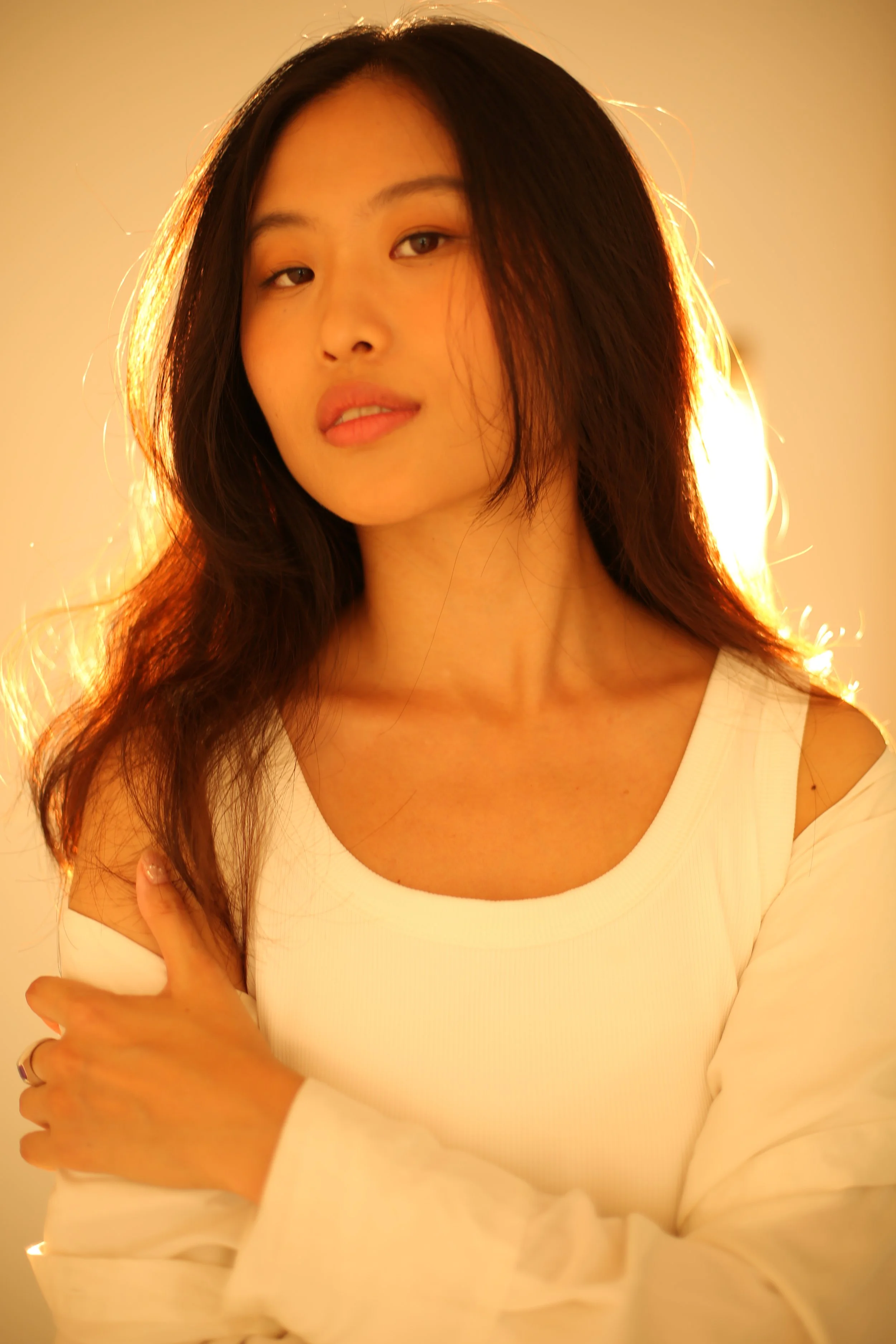 Portrait of a young woman with long dark hair in warm lighting, wearing a white top and slightly tilting her head with a relaxed expression.