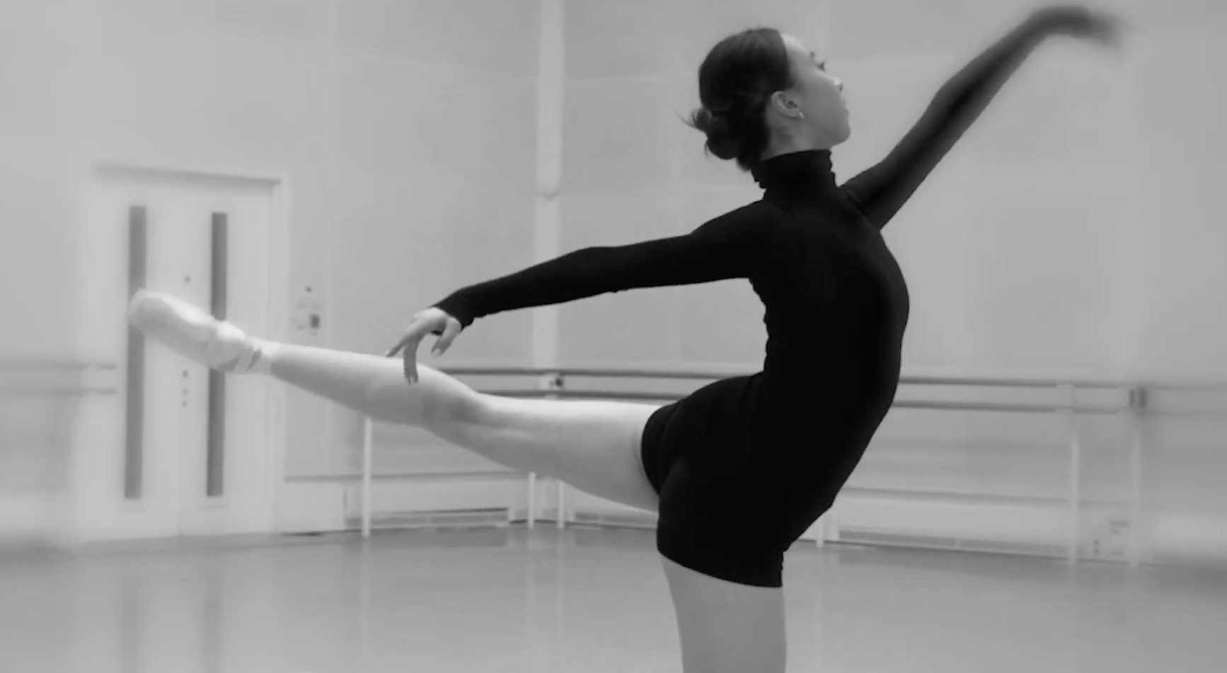 A ballerina in a black outfit practicing dance at a ballet studio, holding a barre with one hand and extending the other arm forward.