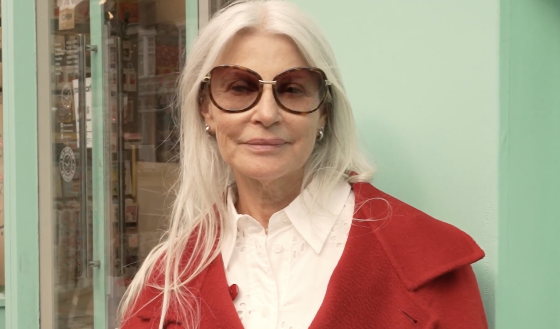 An older woman with long white hair wearing dark sunglasses, gold earrings, a white blouse, and a red coat, standing outside near a store with glass doors and a pastel green wall.