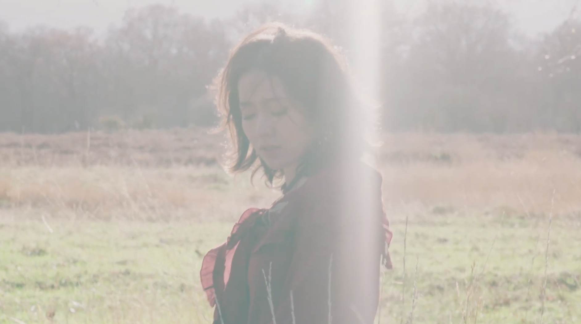 A woman with shoulder-length dark hair, wearing a dark jacket and carrying a red backpack, standing in a grassy field during daytime with sunlight creating a bright, hazy effect.