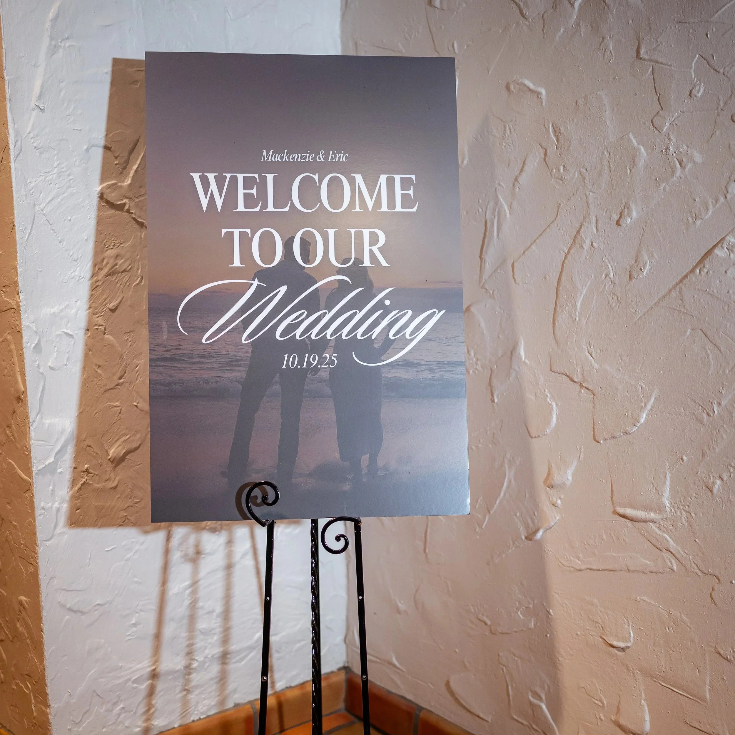 A wedding reception sign with a silhouette of a couple holding hands on the beach at sunset, welcoming guests to Mackenzie and Eric's wedding on October 19, 2025.