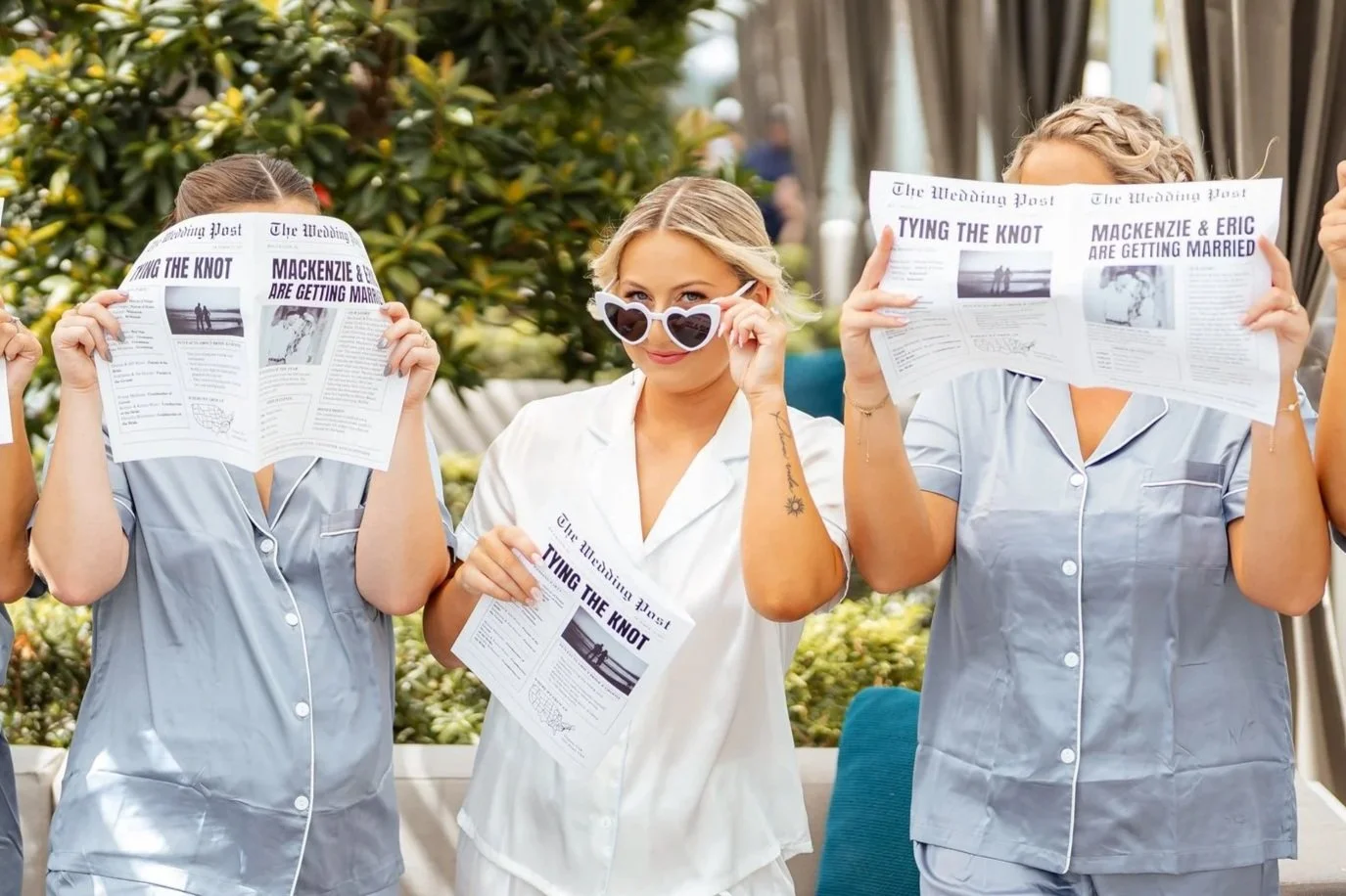 Three women in pajamas, two holding newspapers and one wearing sunglasses, seated outdoors with greenery in the background.