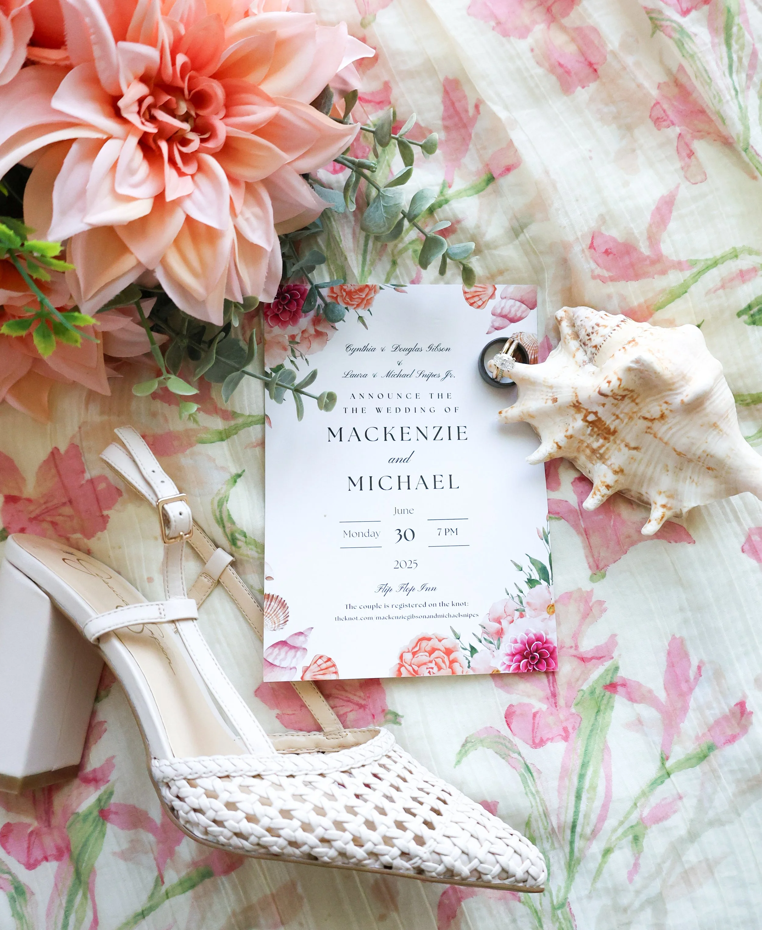 A wedding invitation surrounded by pink flowers, a white seashell, a white woven heeled shoe, and a seashell, laid on a floral tablecloth.