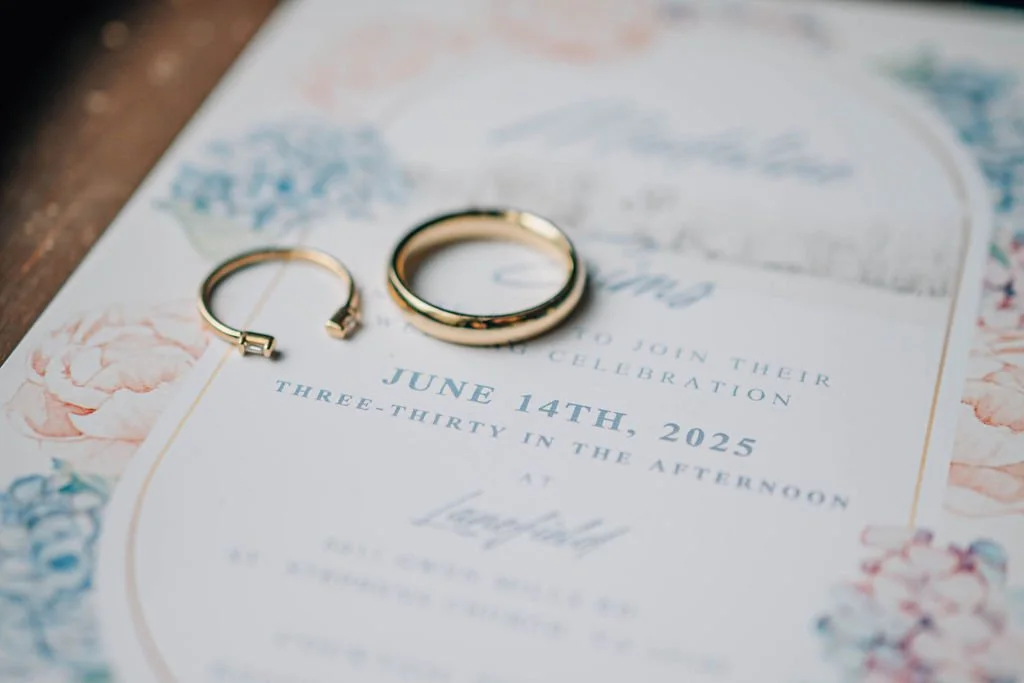 Wedding invitation with two gold rings placed on top, surrounded by a border of pink and blue flowers.