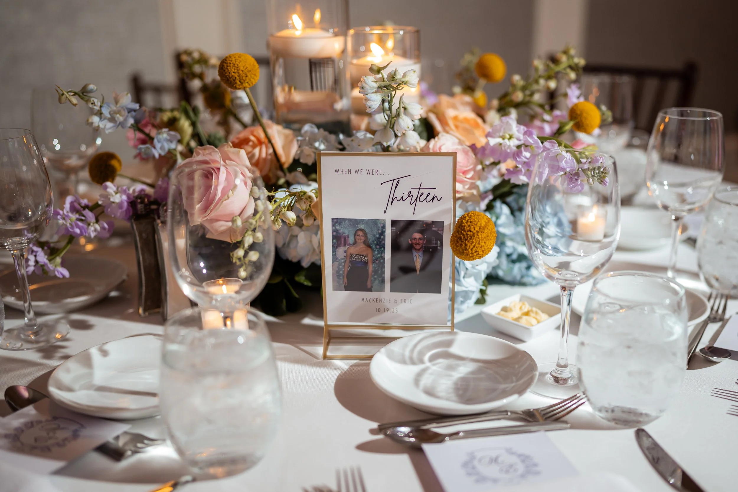 A decorated dining table with floral centerpieces, candles, wine glasses, plates, silverware, and a framed photo card with the names Mackenzie and Eric, for a wedding or special celebration.