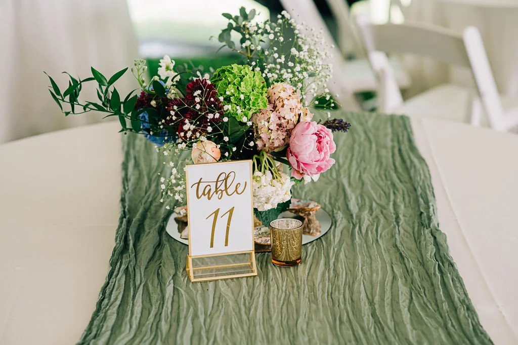 A floral centerpiece on a wedding table with a table number sign reading 'table 11', a small glass candle holder, and a green table runner.