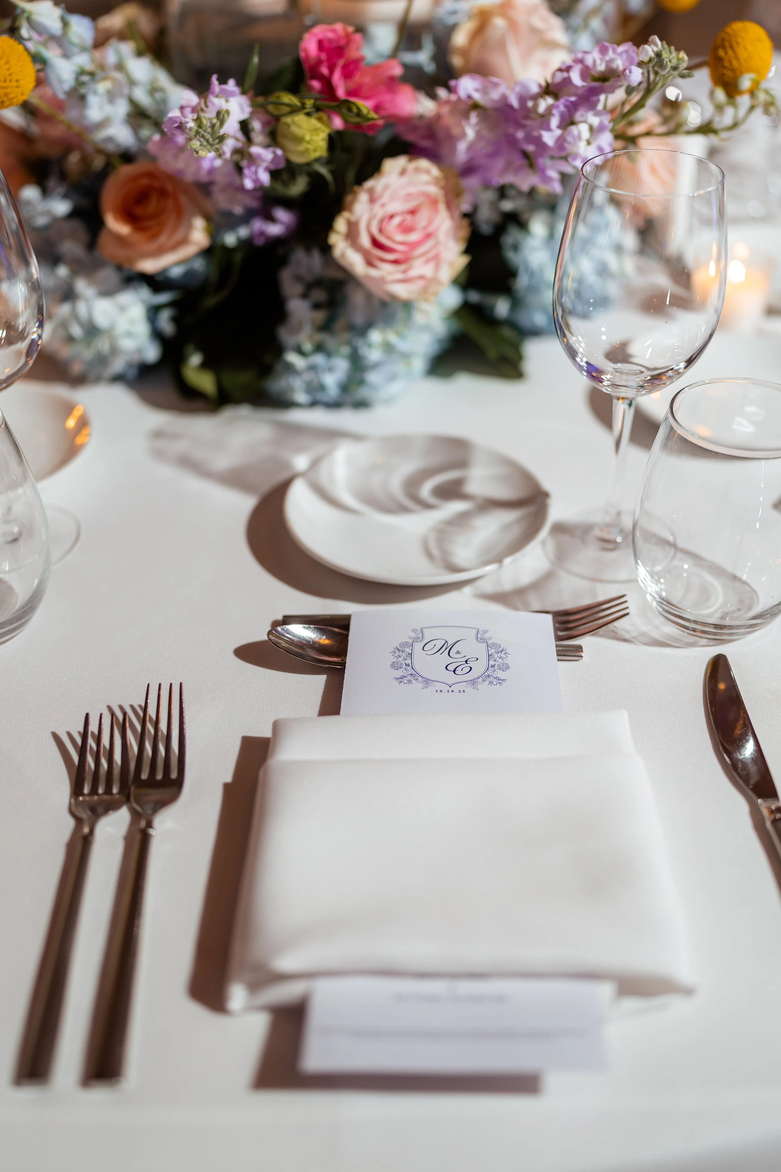 A table setting with silverware, a folded white napkin, and a place card with decorative lettering that says "Mr & Mrs" along with a wedding date. In the background, there is a floral centerpiece with pink, purple, white, and yellow flowers.