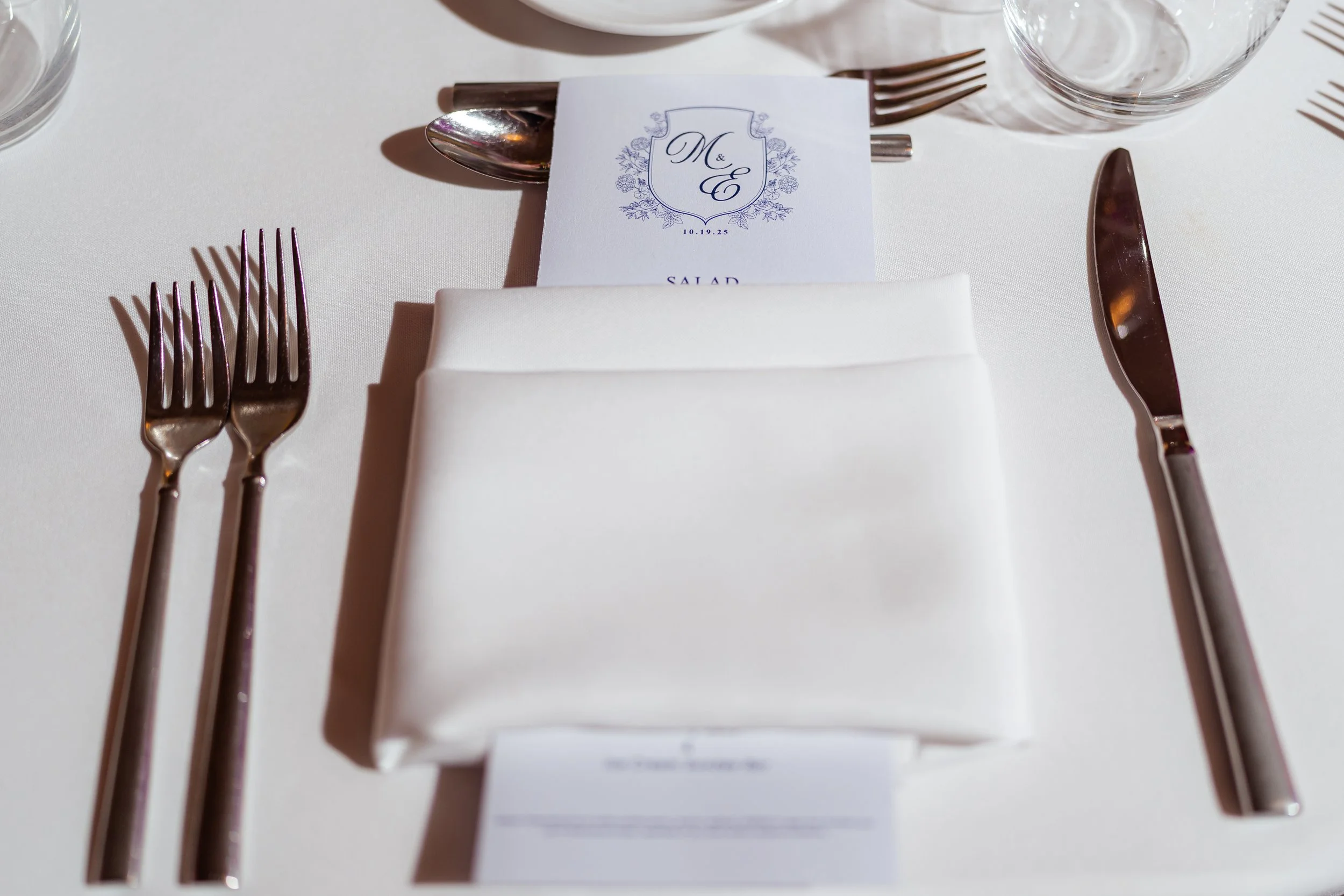 Setting for a formal dining table with silverware, a white napkin, a printed menu, and a glass, with soft lighting.