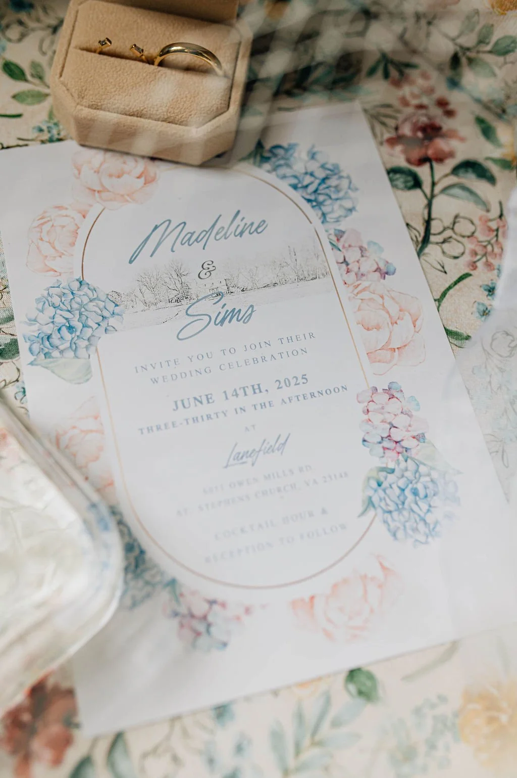 Wedding invitation featuring floral design with blue and pink hydrangeas, gold accents, and text with the names Madeline and Sima, date June 14th, 2025, and location details.