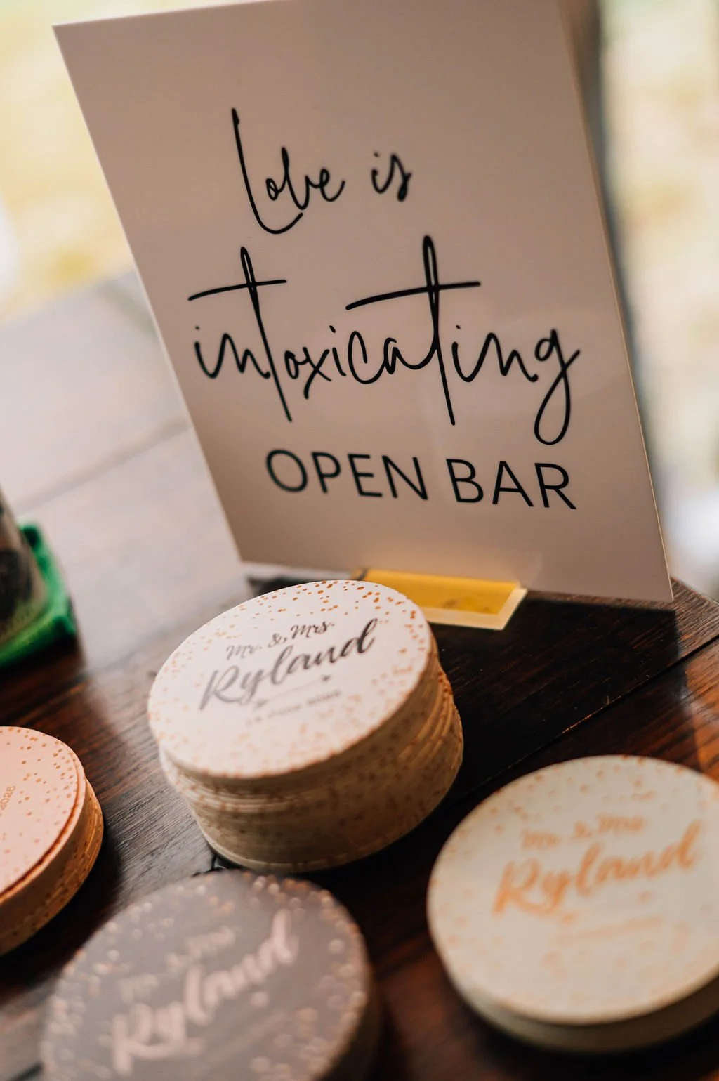 A sign on a table reads "Love is intoxicating OPEN BAR". There are stacks of round coasters with white and gold designs and the name "Mr. & Mrs. Ryland" printed on them.