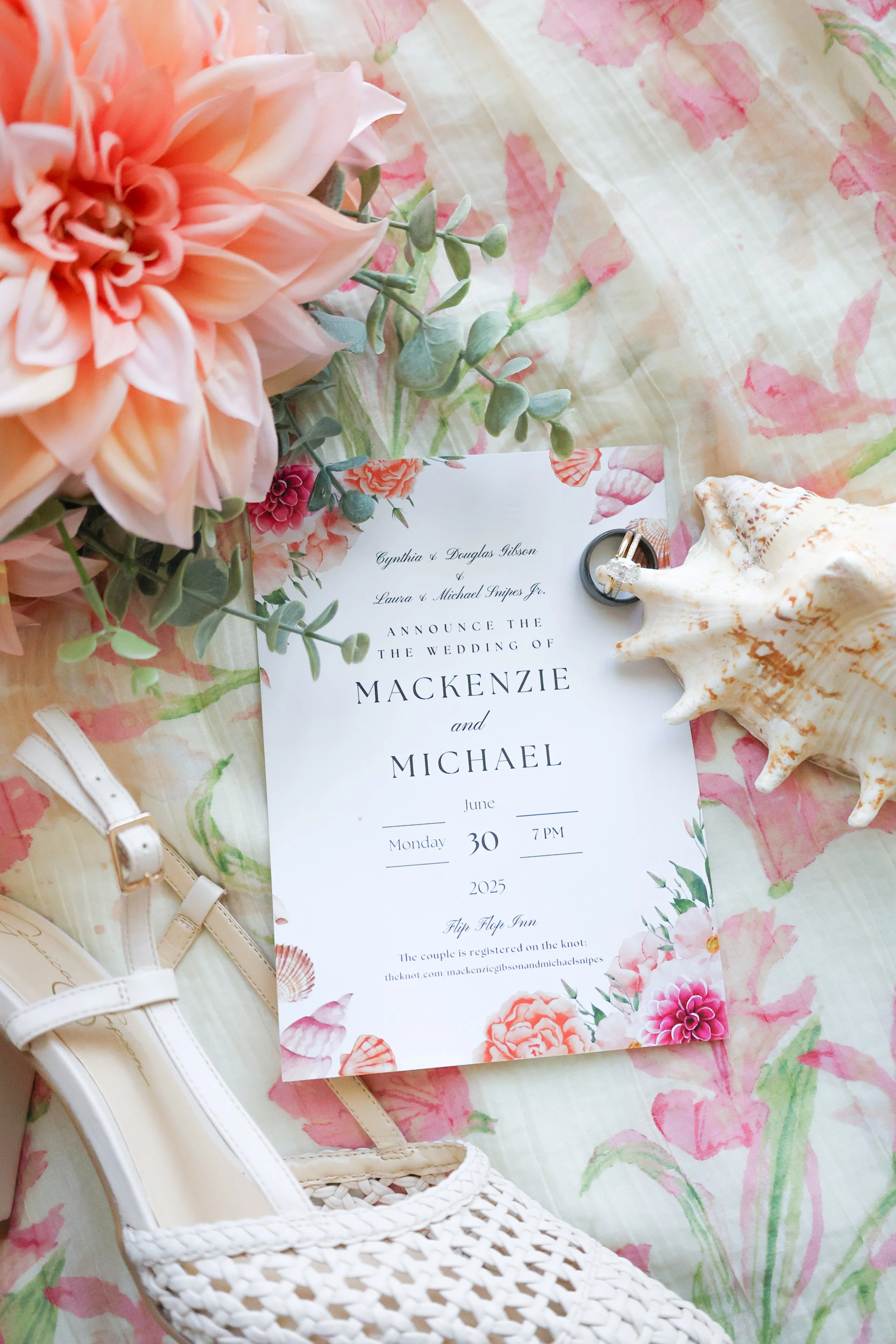 Wedding invitation with floral border, surrounded by a pink flower, shell, and white high-heeled shoes on a floral fabric background.