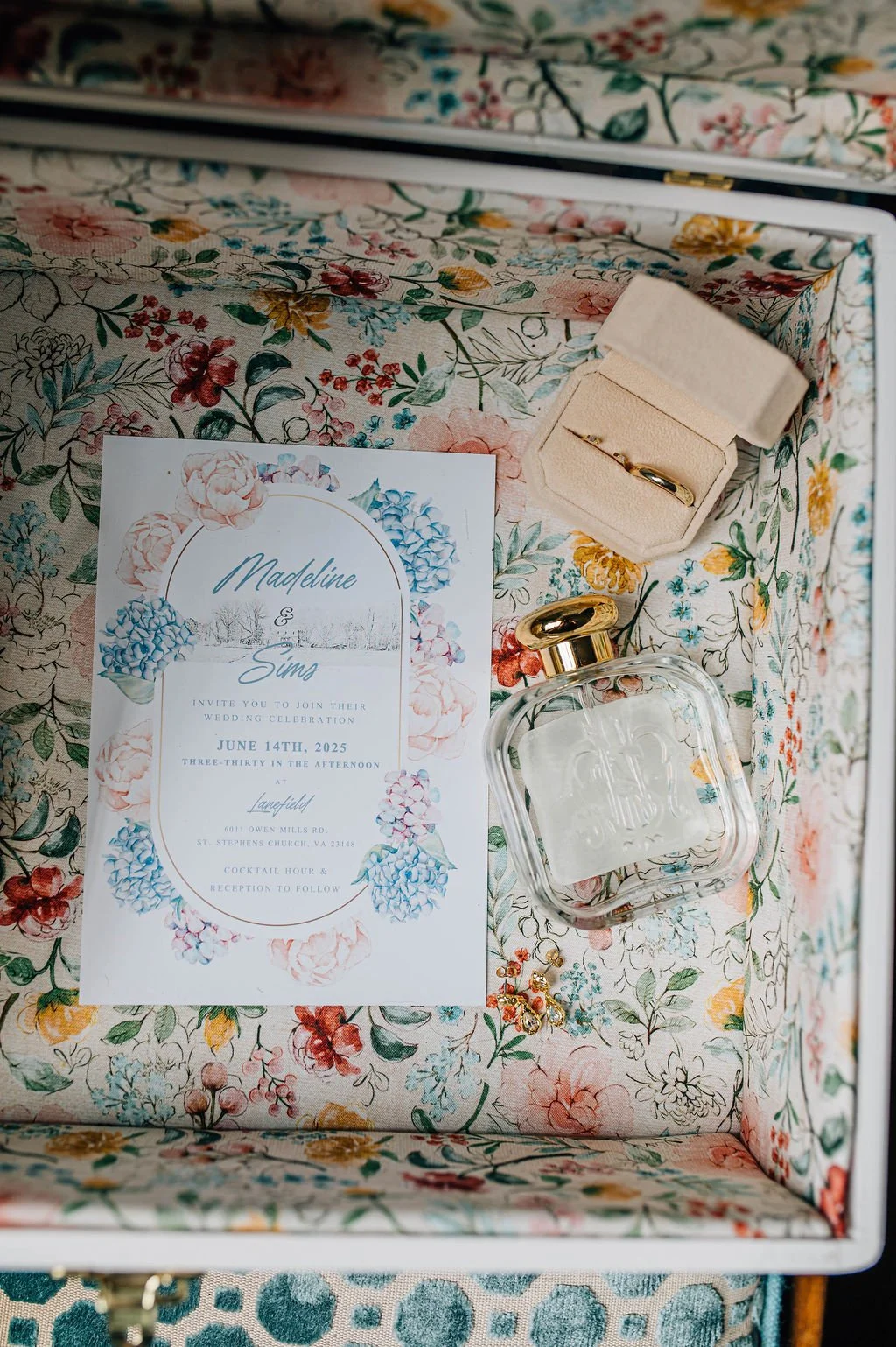 A floral wedding invitation card, a small jewelry box, a perfume bottle, and earrings are arranged inside a box with floral patterned lining.