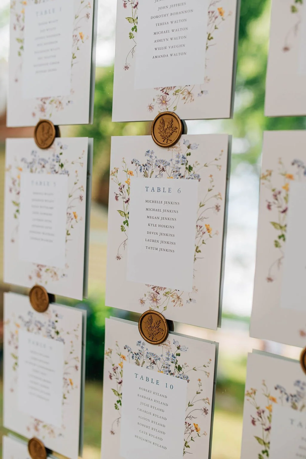 Wedding seating chart with floral border design, showing tables with names of guests, held with wax seals, set in front of a window with greenery outside.