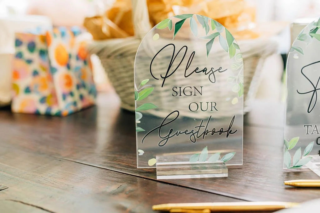 A clear acrylic sign with the words "Please sign our guestbook" written in black cursive and print, decorated with green leaves, placed on a wooden table. In the background are gift bags and a wicker basket.