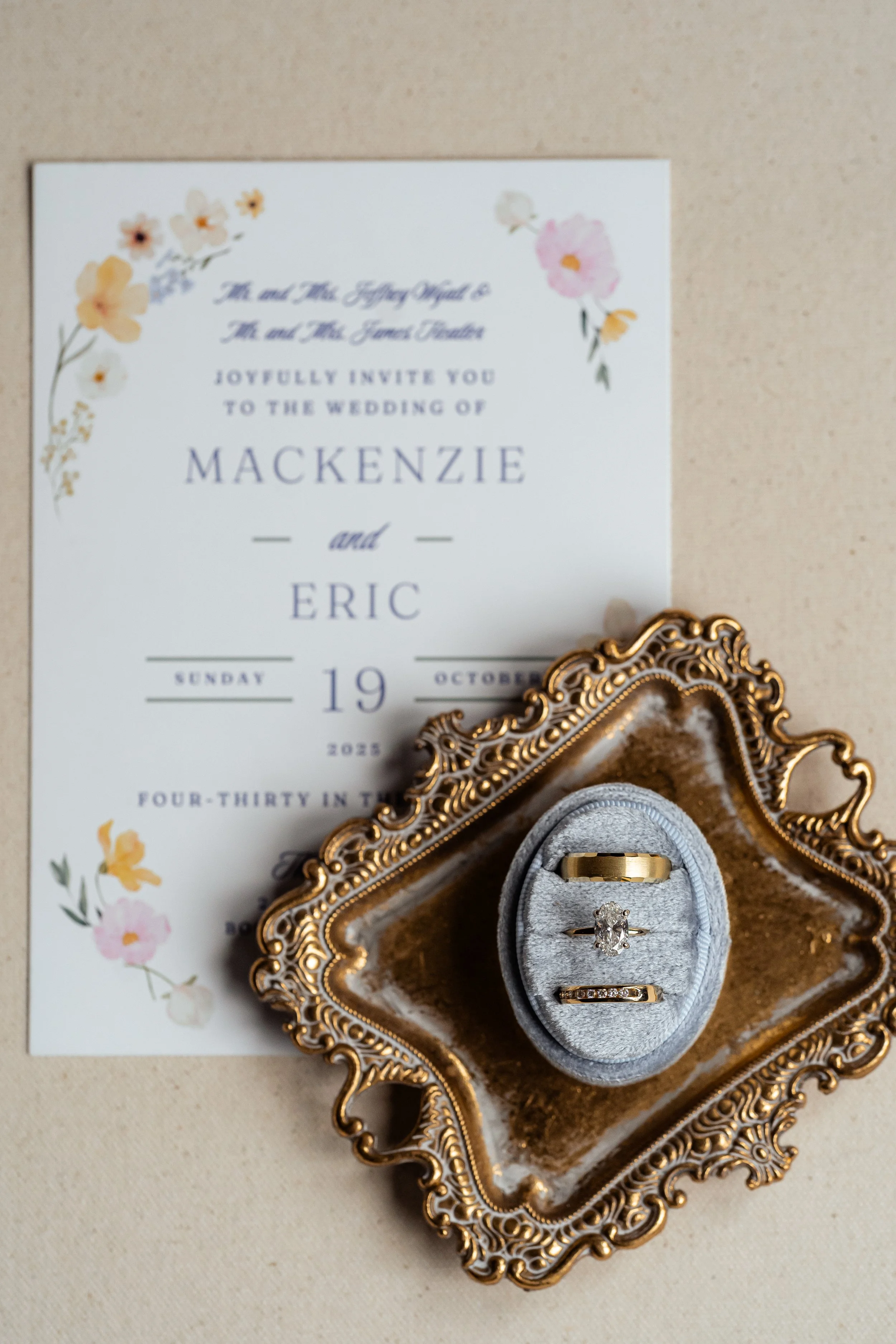 Wedding invitation with floral design, wedding rings in a gray velvet ring box on an ornate gold tray.