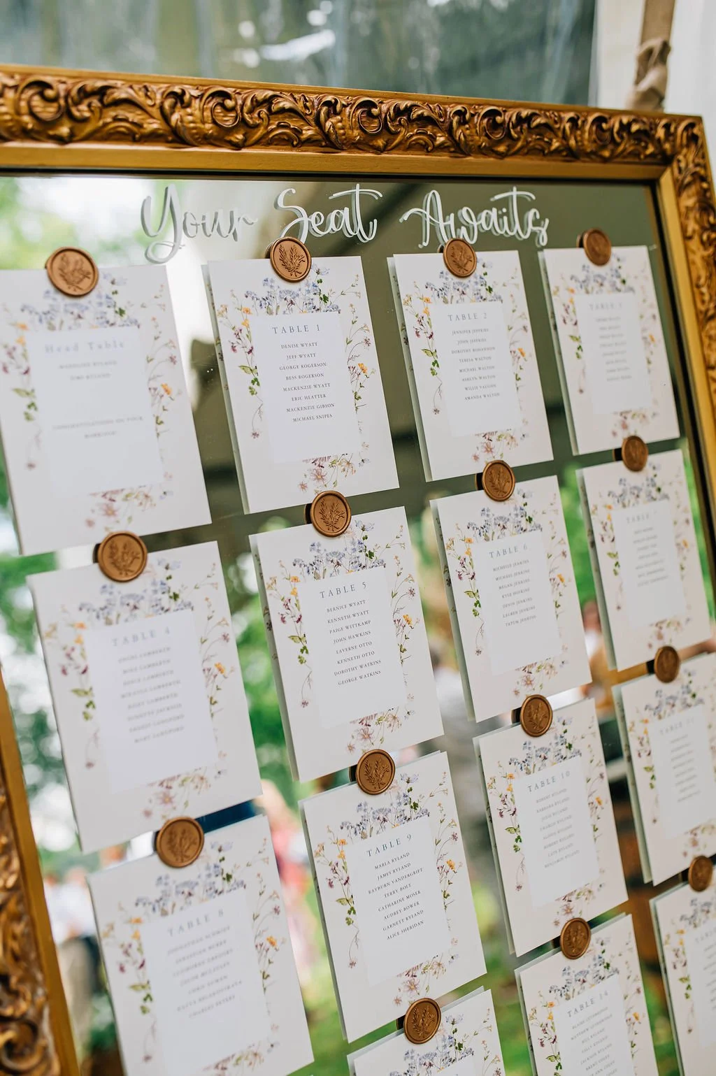 A decorative wedding seating chart with floral designs, pinned with wax seals, displayed on an ornate gold-framed mirror