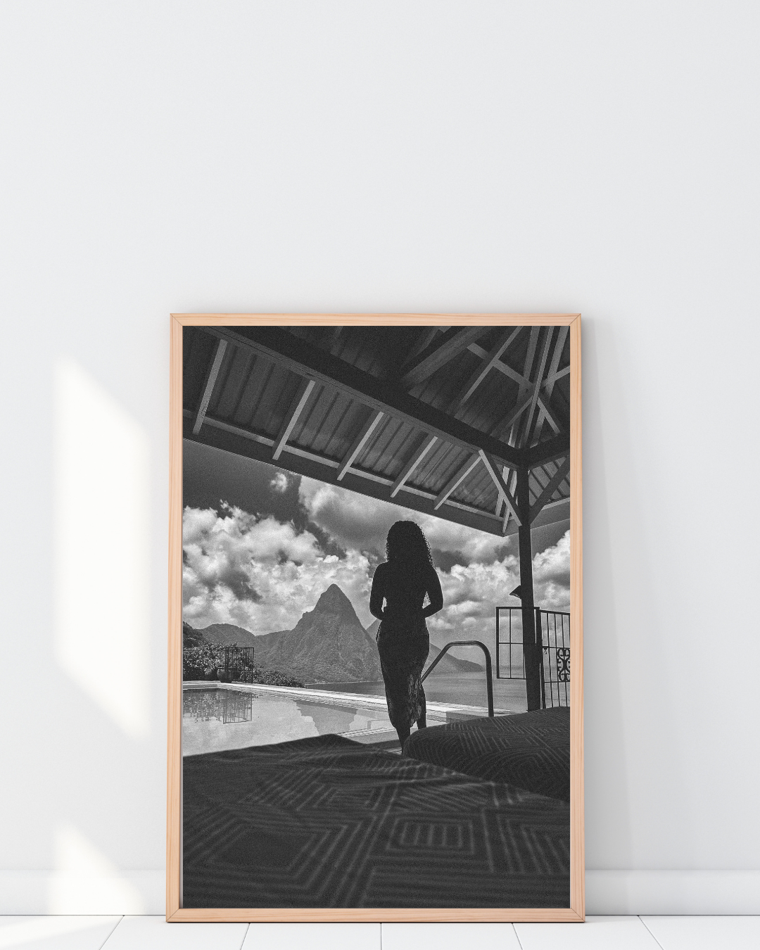 A black-and-white photo of a woman standing near a pool, gazing at the mountains and clouds in the distance, under a roofed structure.