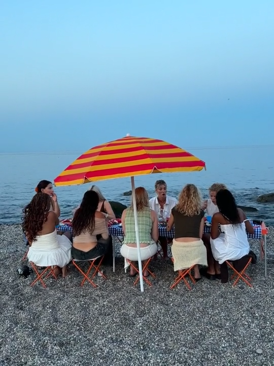 Seven women sitting at a picnic table under a large red and yellow striped umbrella on a beach, with the ocean and sky in the background.