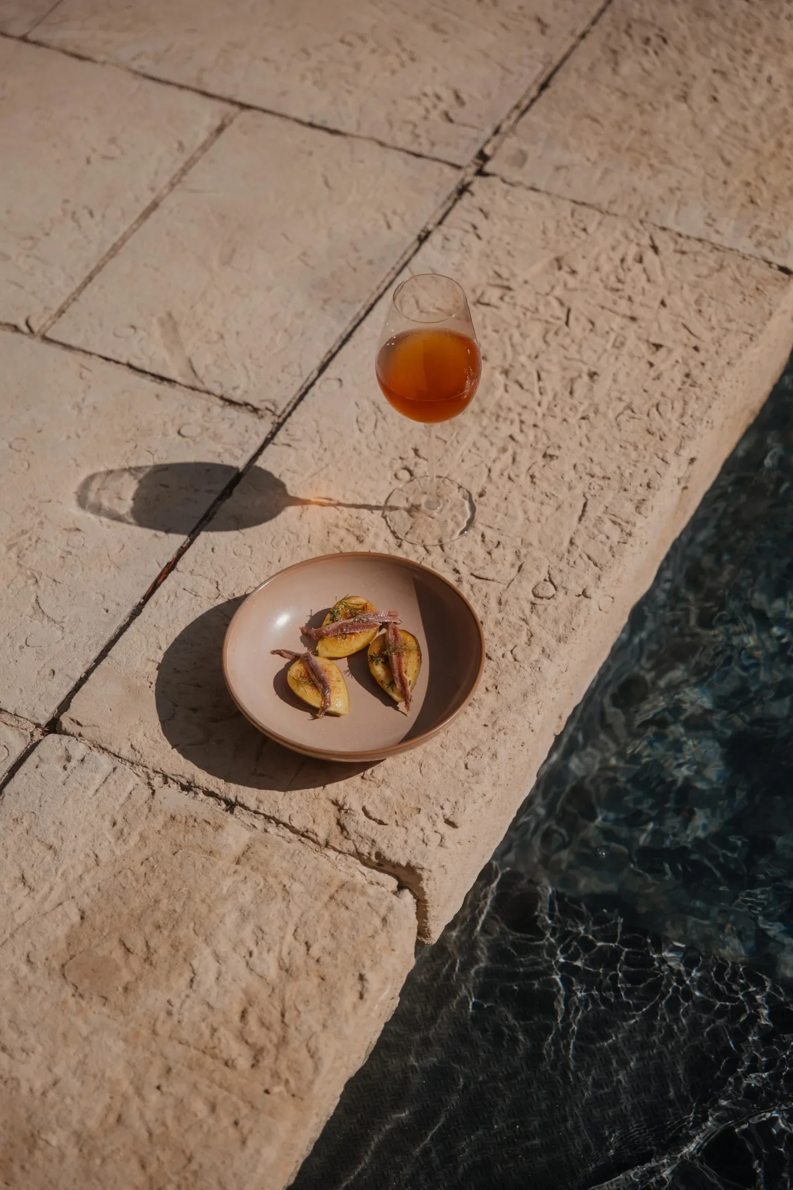 A glass of amber-colored drink and a small plate with halved lemons topped with anchovies on a stone ledge beside water.