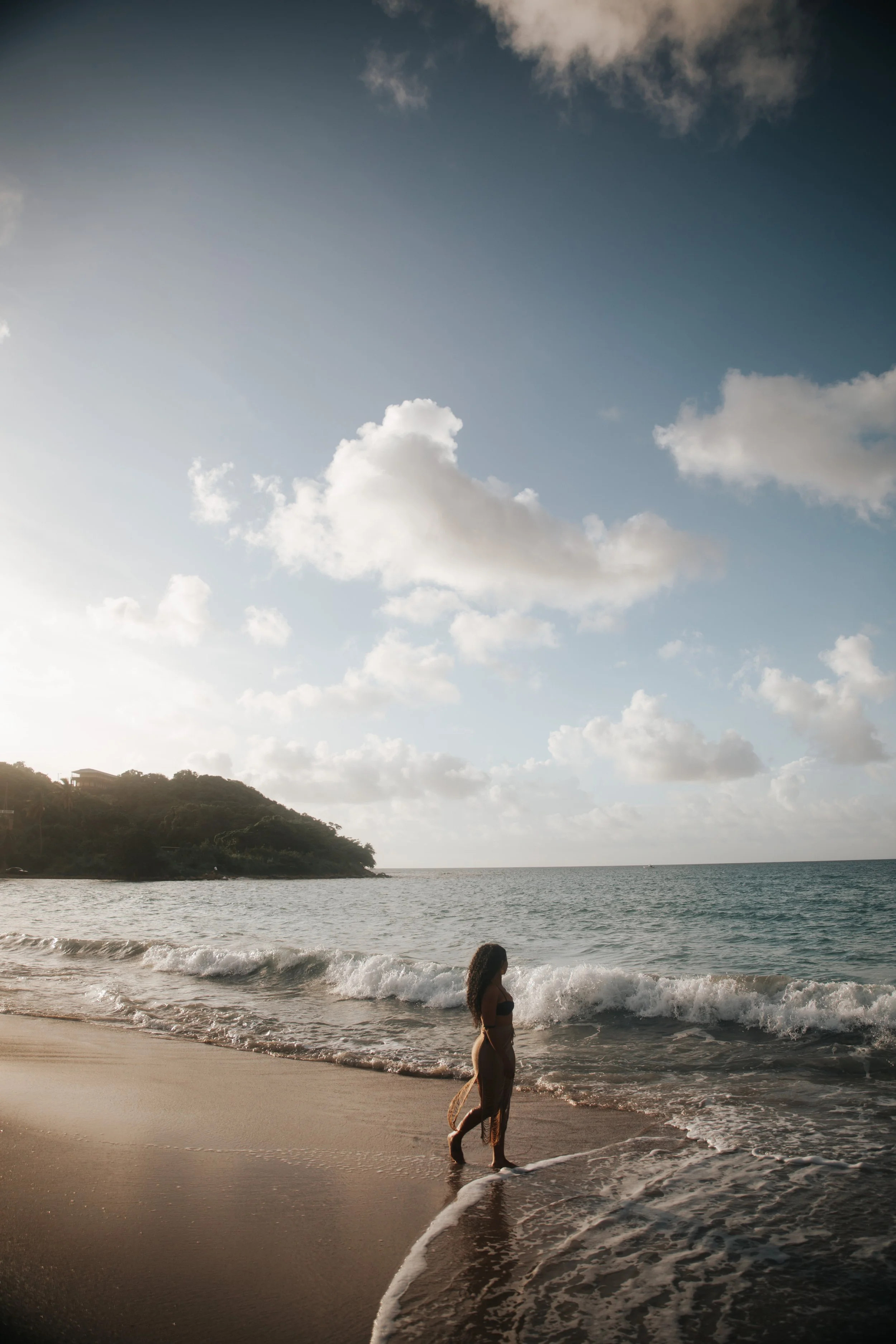 A woman in a black bikini walking along the shoreline on a beach with gentle waves, a green hill in the distance, and a partly cloudy sky.