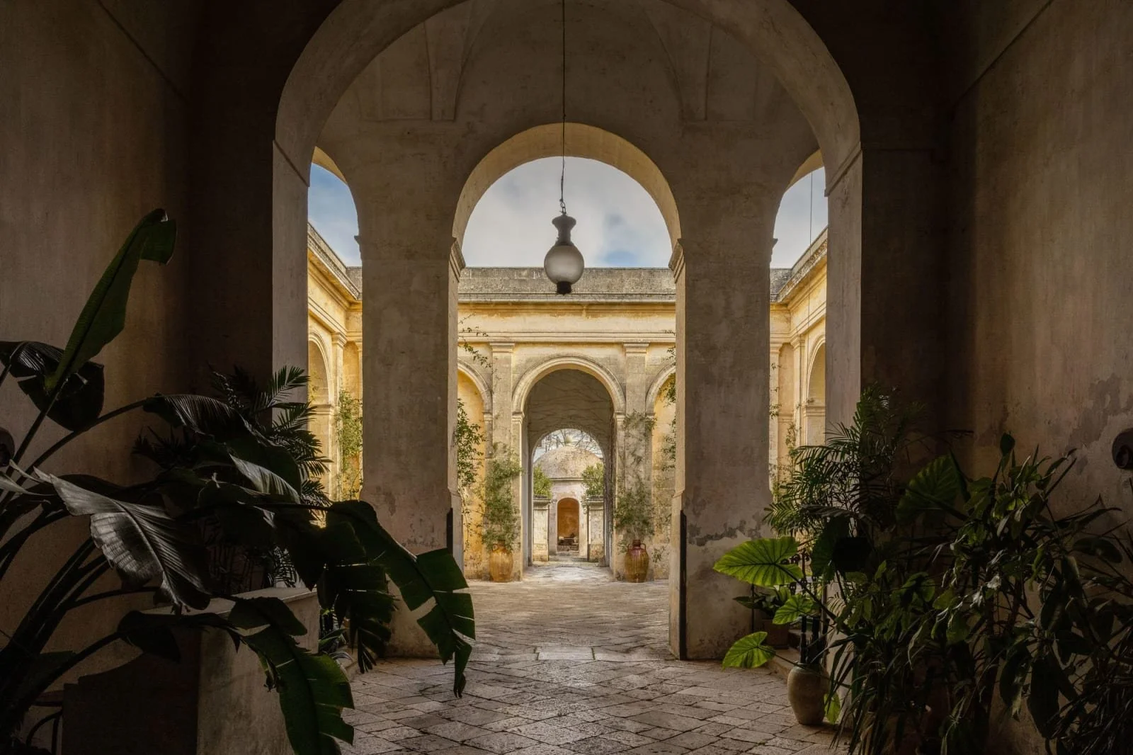 View through arched doorways of a historic courtyard with potted plants, stone walls, and a cloudy sky.