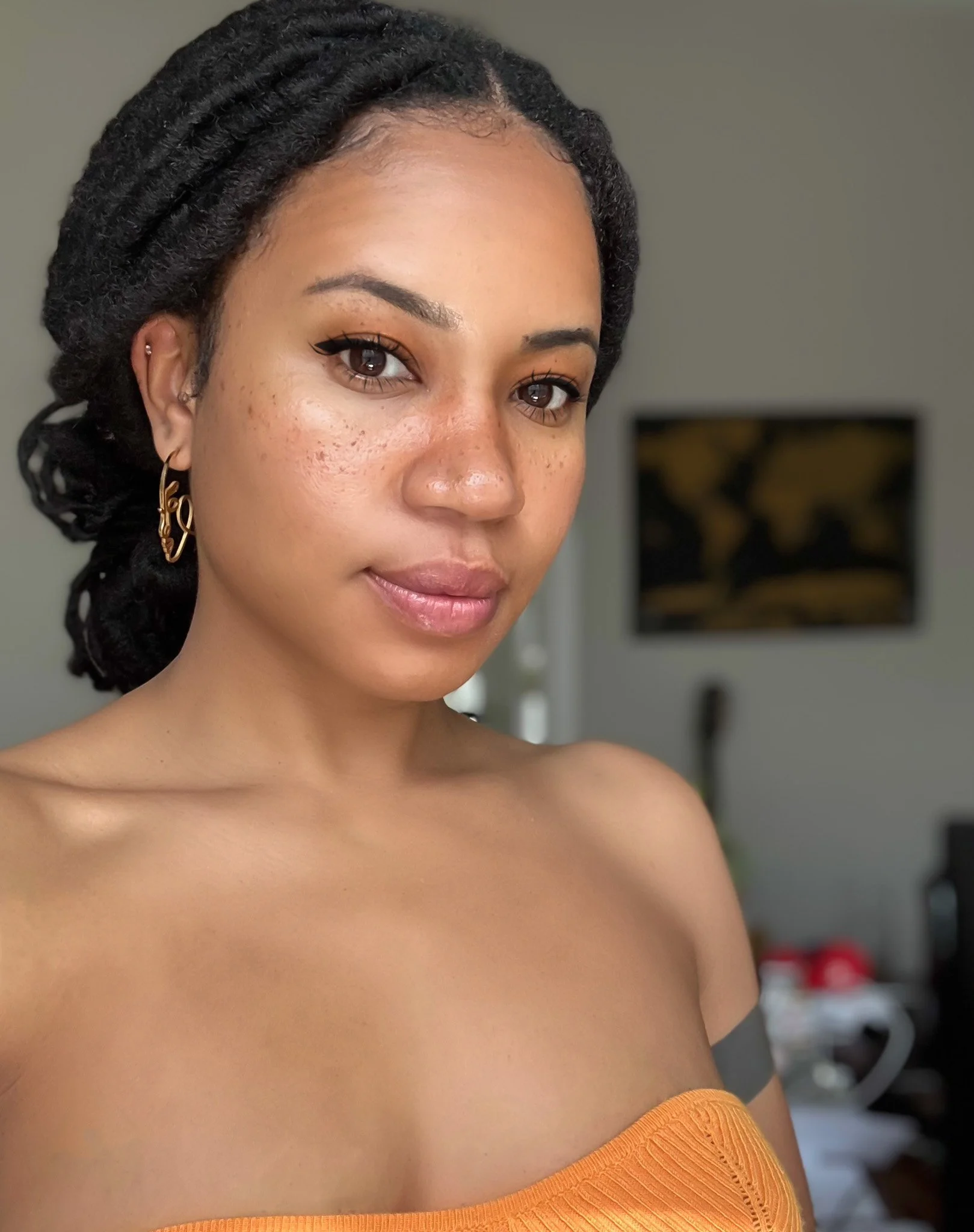 Close-up of a young woman with medium brown skin, dark braided hair, and gold earrings, wearing a strapless orange top. She's in a room with a blurred background including a wall-mounted TV and some furniture.