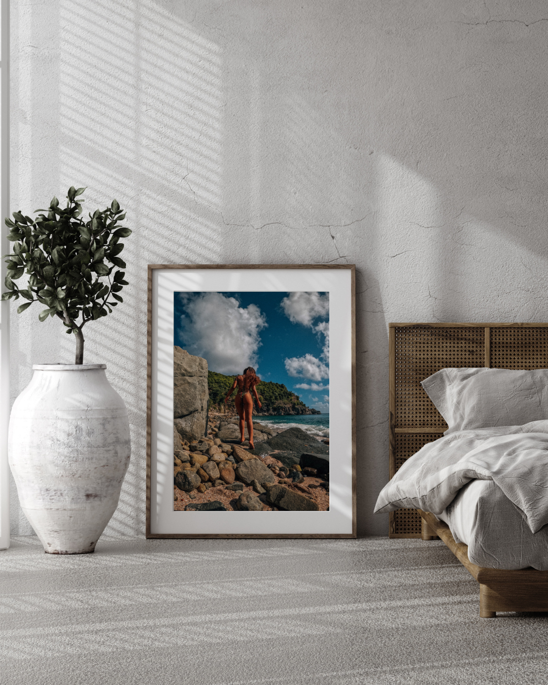 A framed photograph of a woman on a rocky beach, with a partly cloudy sky and lush green hills in the background, leaning against a textured gray wall in a bedroom with a large white vase and green plant to the left, and a bed with a woven headboard 