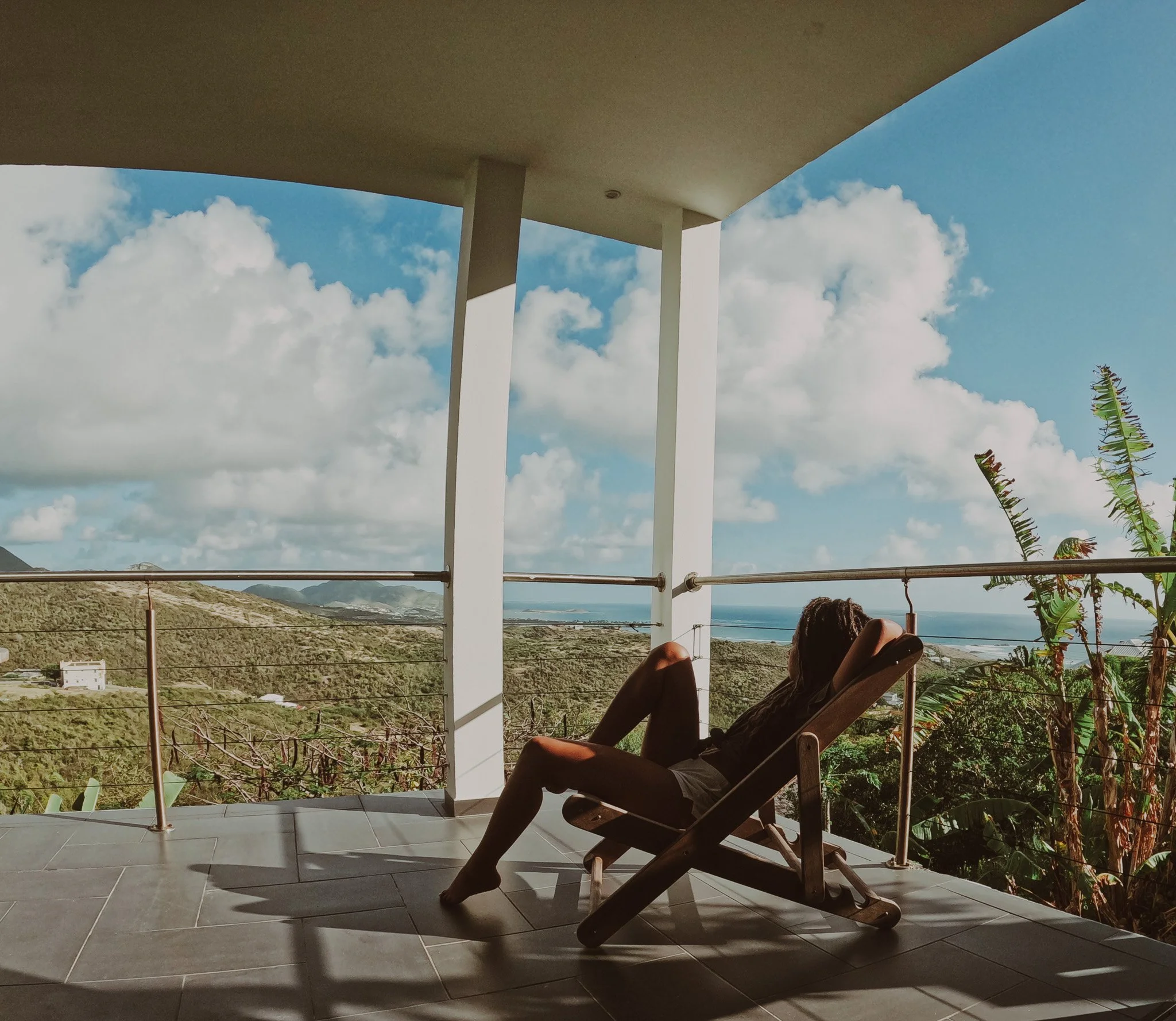 Person reclining on a lounge chair on a balcony, overlooking a lush green valley with hills, trees, and a distant ocean under a partly cloudy sky.